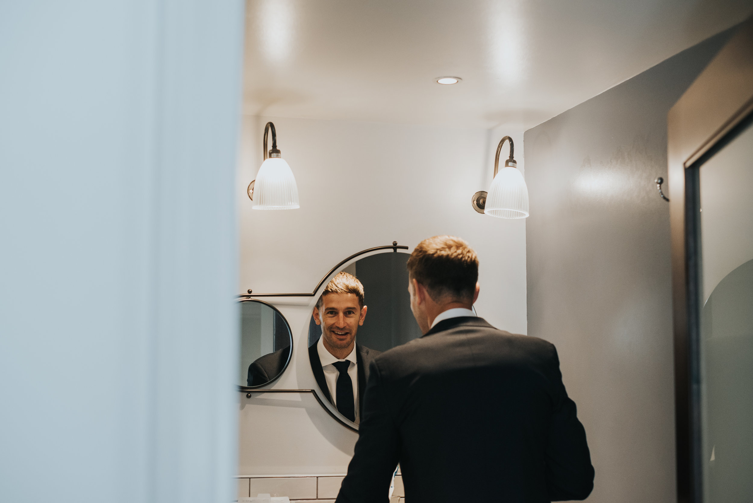 The groom catches the photographer snapping him in the bathroom mirror.