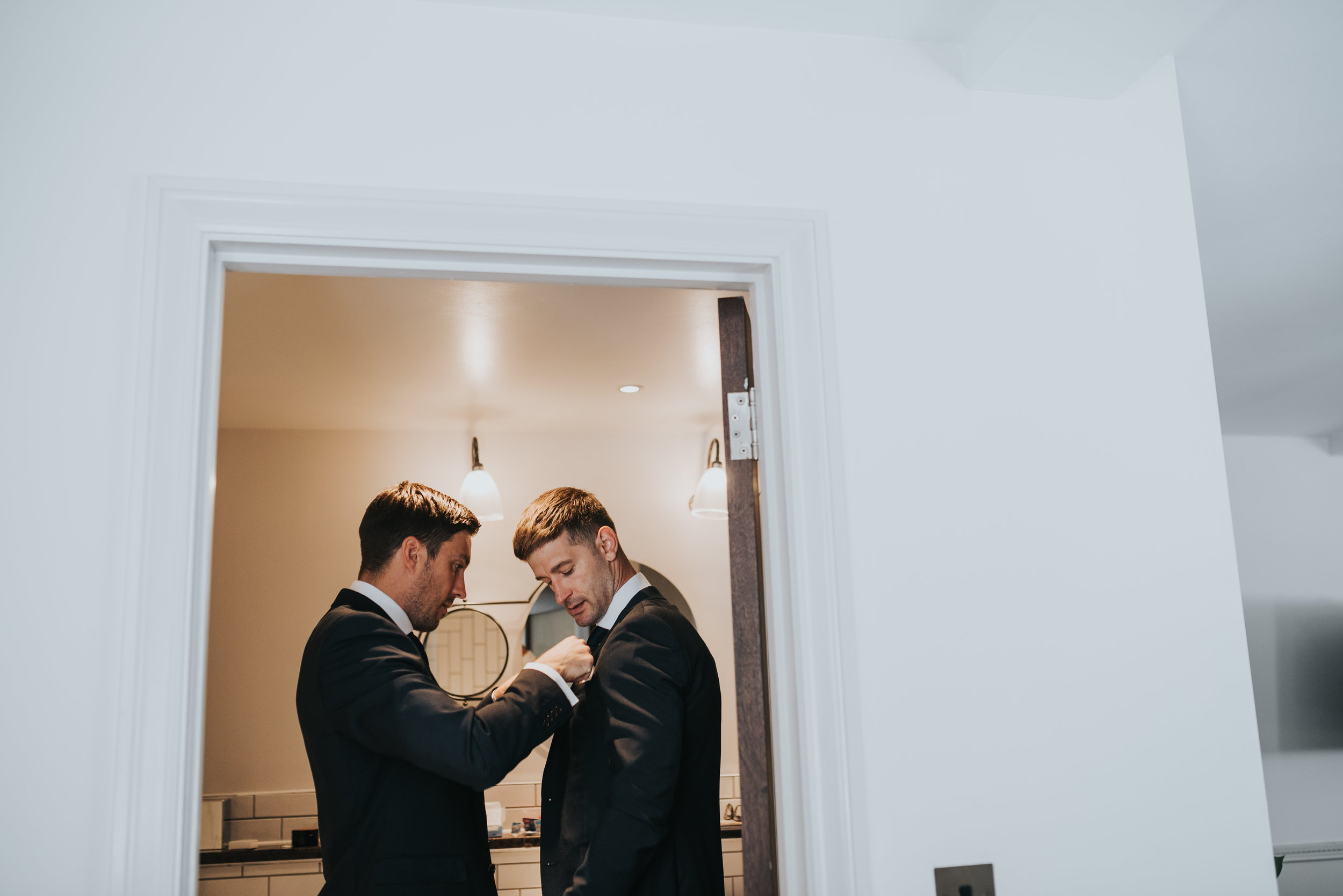 The groom has his groomsman fix his suit for him. 