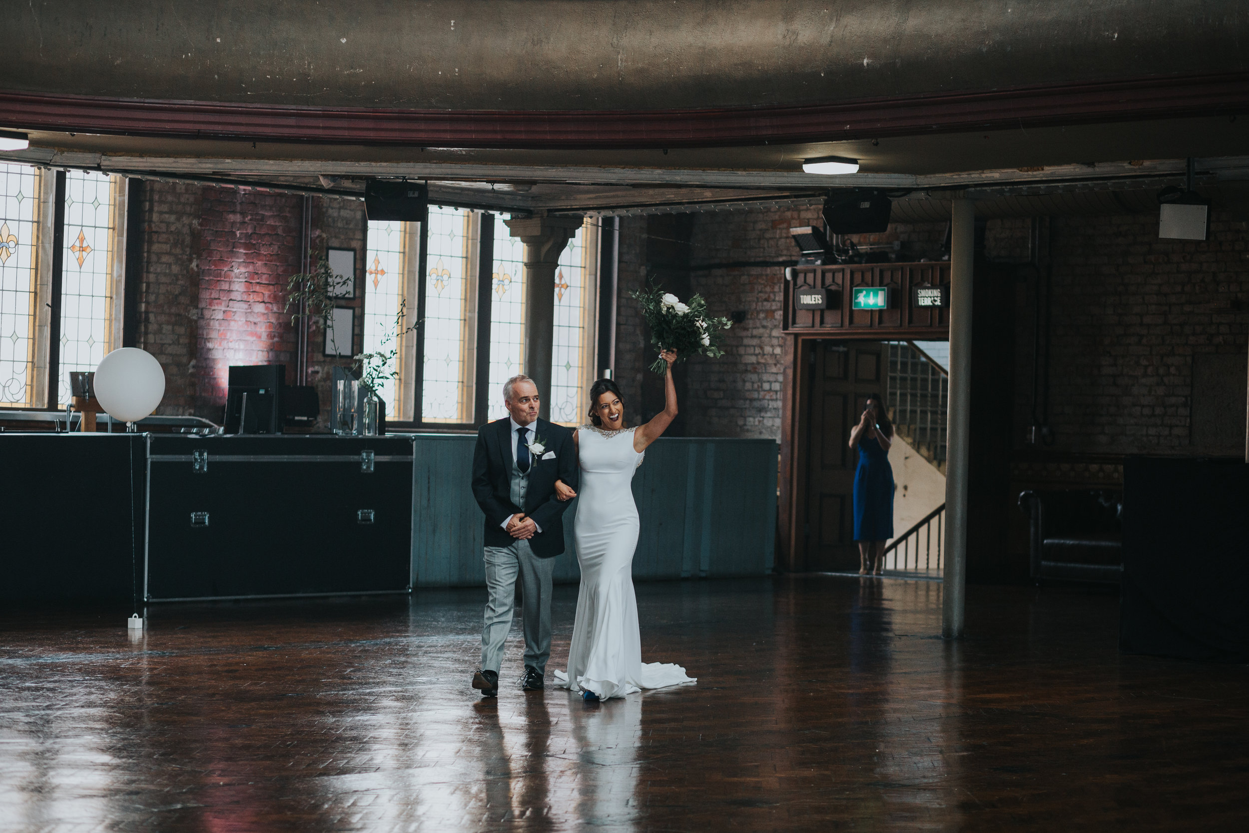 The Bride raises her flowers up to a cheer from her wedding guests as she enters the hall with her father on her arm. 