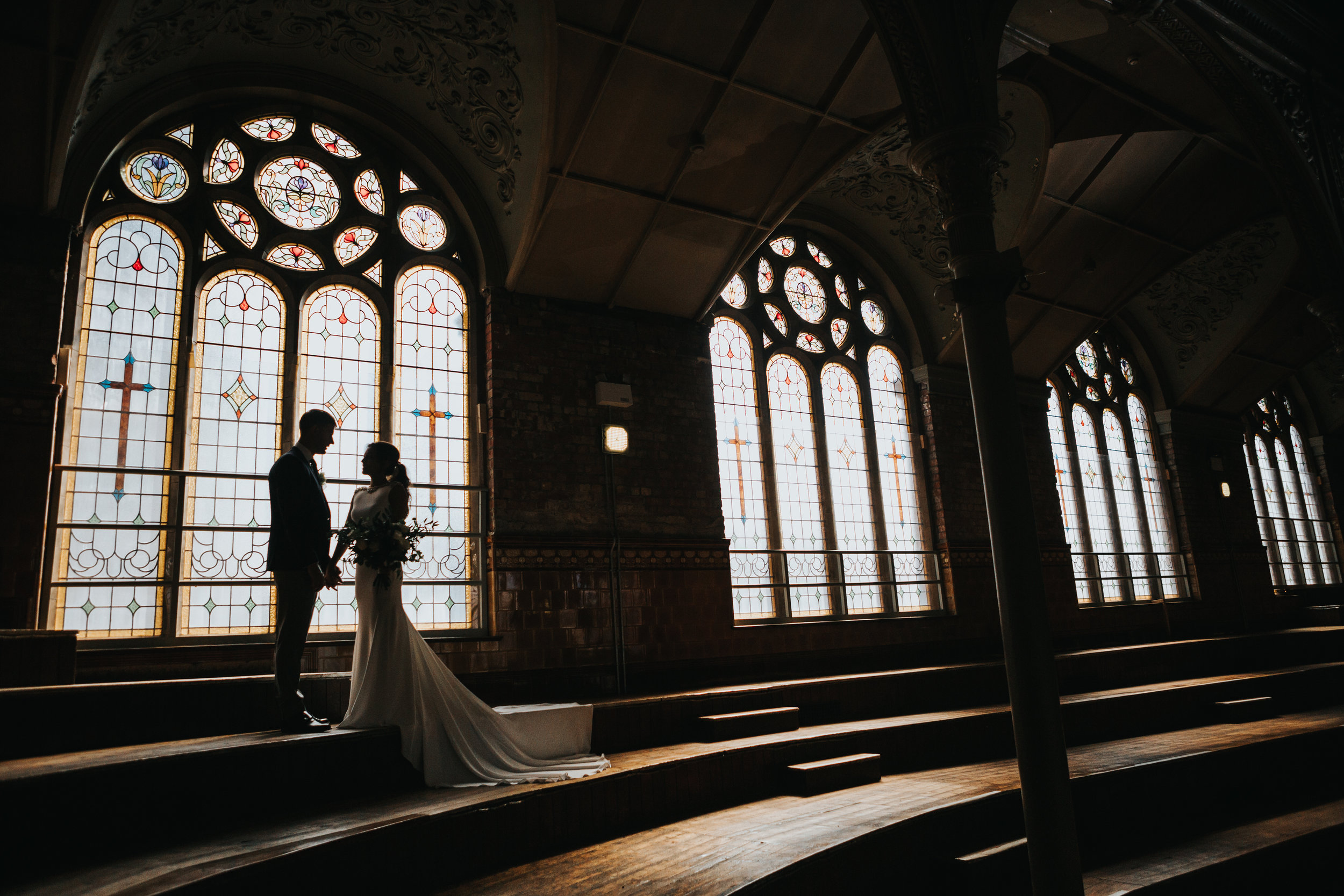 The couple stand holding hands in front of a huge stain glass window. 
