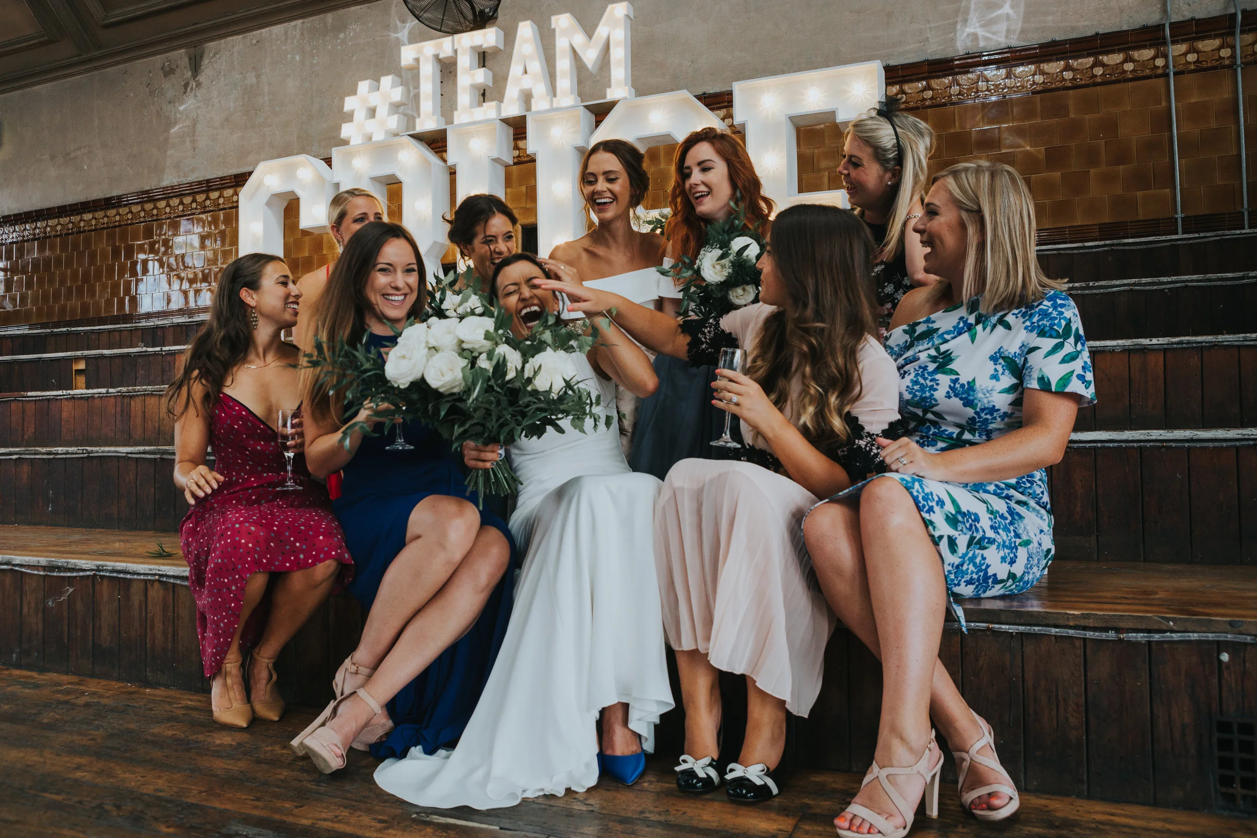 Bride sits in the middle of her female friends laughing as they try and poke her in the face, huge TEAM CRUSE sign behind them. 