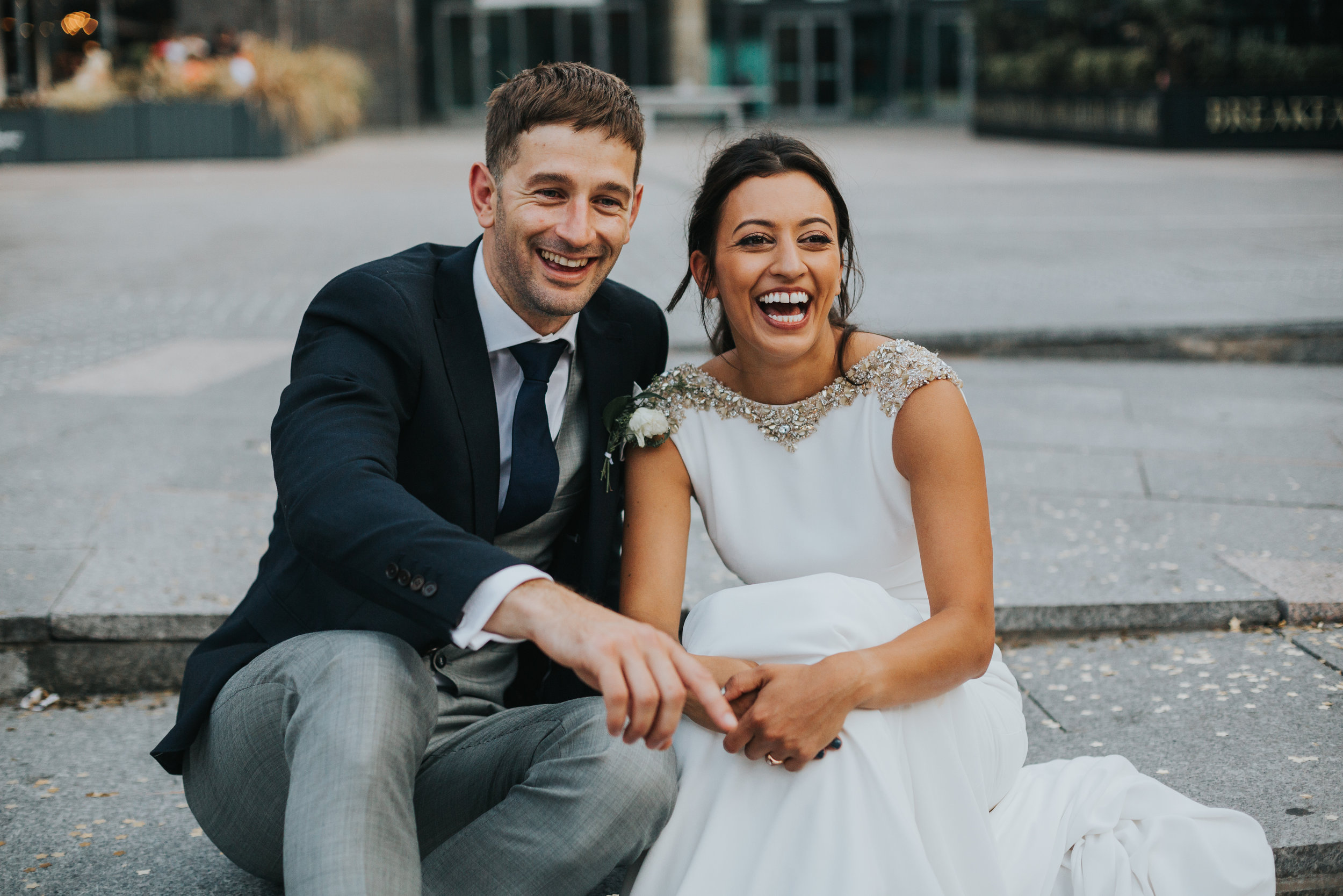 Bride and groom sit laughing together over the road from The Albert Hall Manchester.