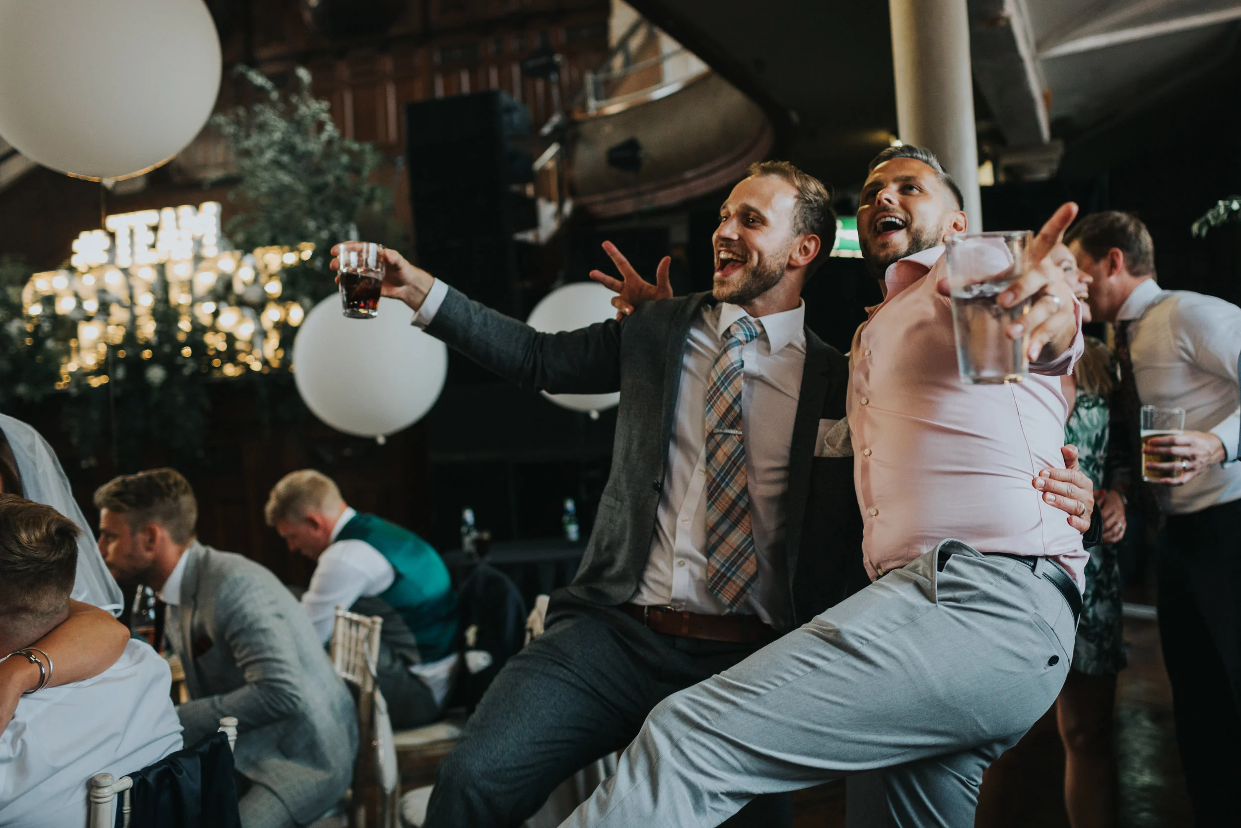 Two male wedding guests dancing with their legs in the air.