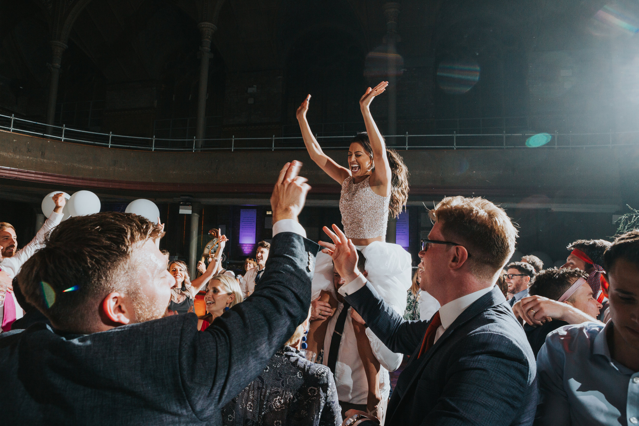 Bride sits on a guests shoulder, her hands in the air singing on her wedding day at The Albert Hall, Manchester. 