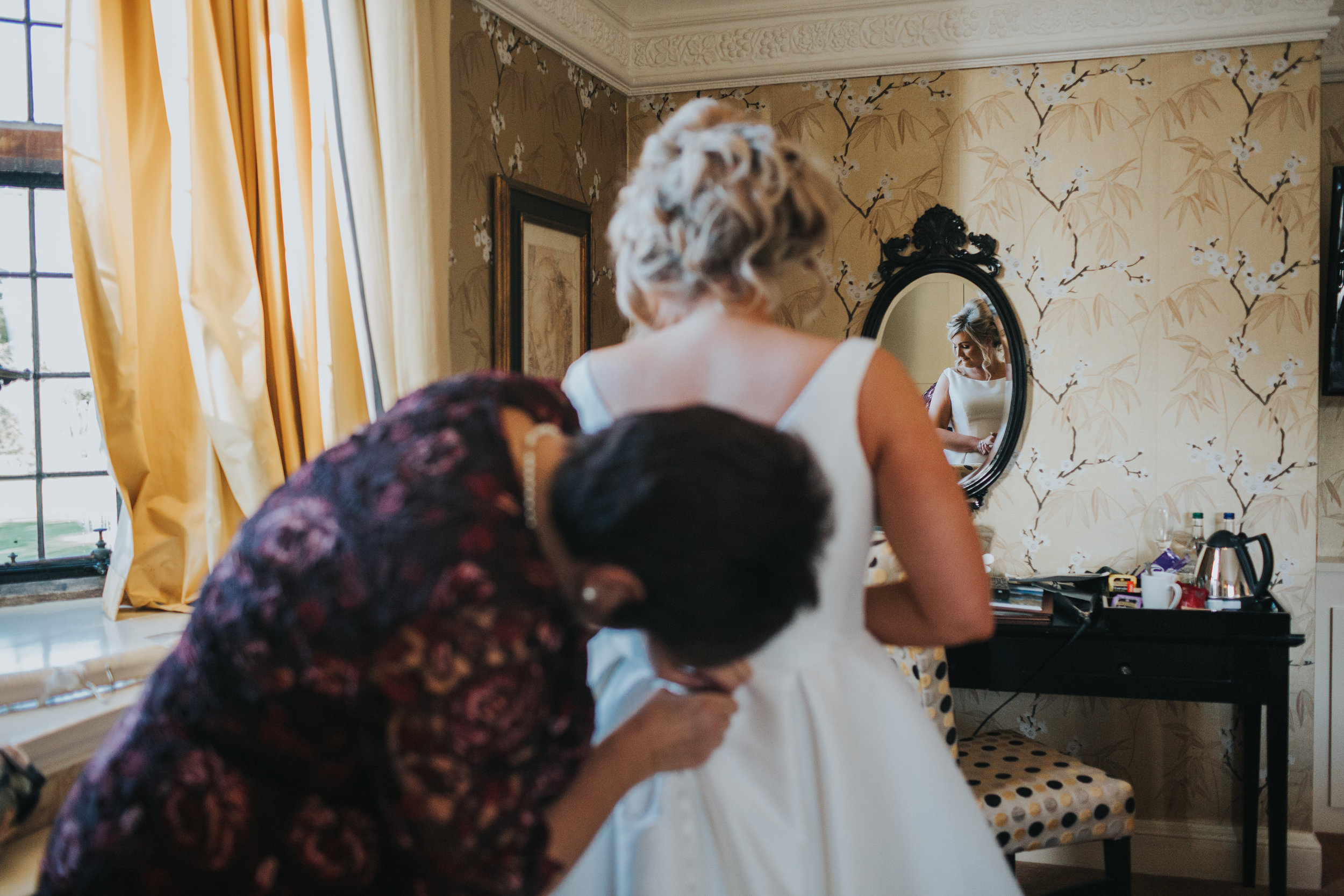 Reflection of bride checking her dress in Snow White Mirror in the background of the shot, while her mum fastens her dress in the foreground. 