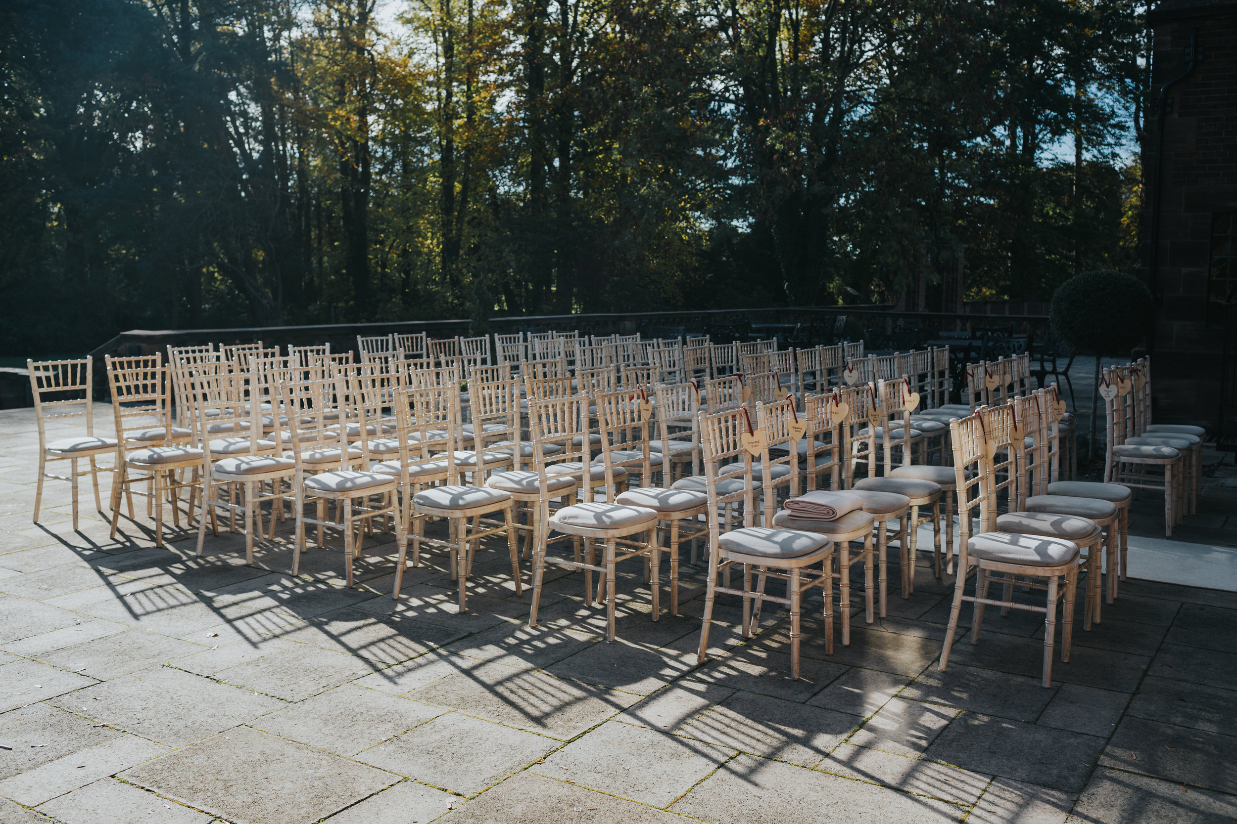 Empty cream wooden wedding chairs, sit empty in the sunlight casting shadows on patio awaiting the wedding guests. 