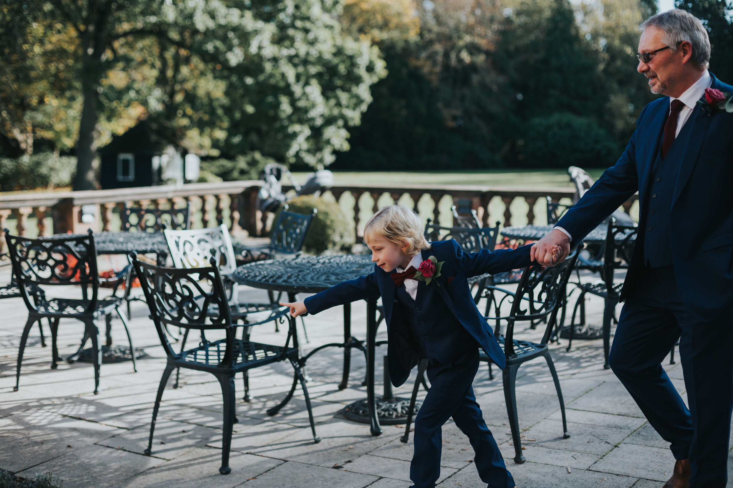 Small boy in a blue suit and red bow tie drags an older groomsmen across the patio. 