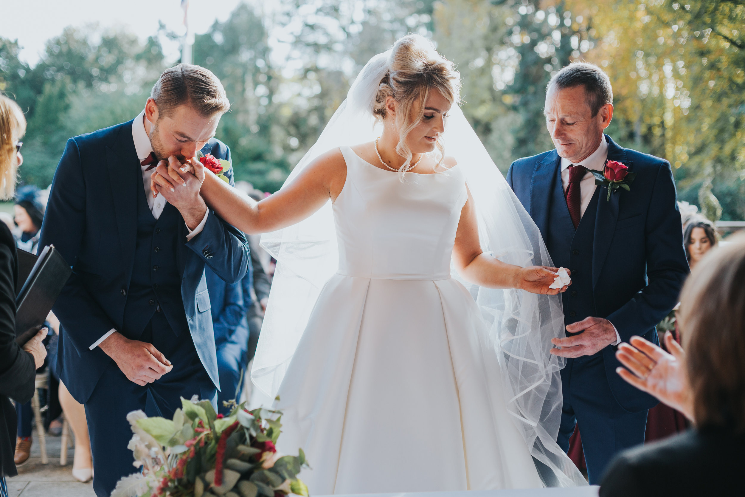 The Groom kisses the brides hand, while her father gives her a tissue to wipe their tears and snot probably.