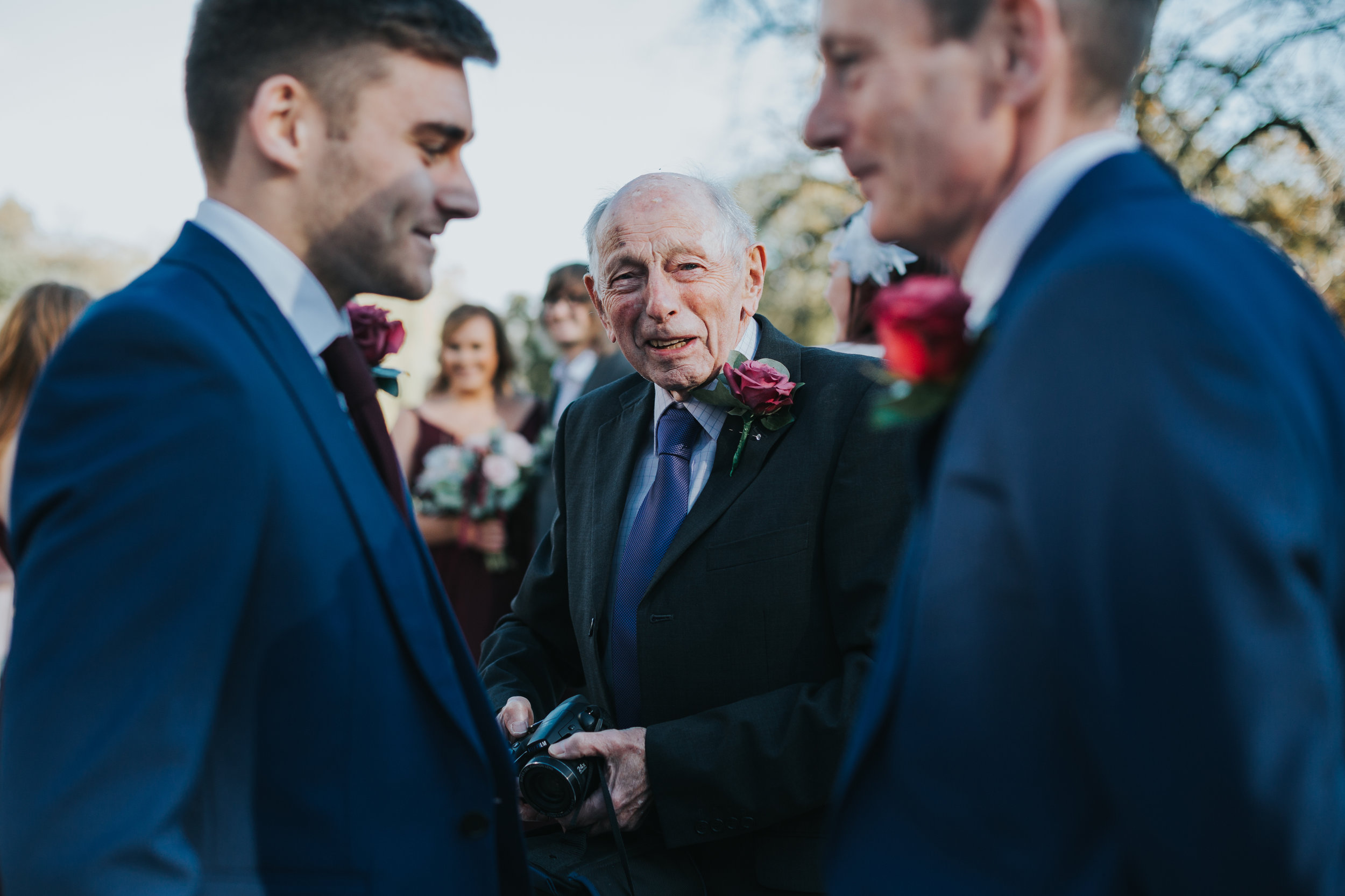 Older male wedding guest holds his camera, framed by two male wedding guests wearing blue. 