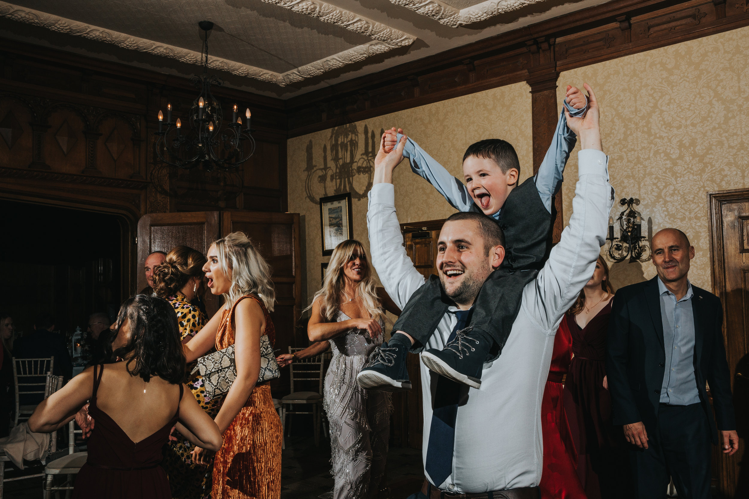 Little boy sits on his dads shoulders cheering as wedding guests dance around them.
