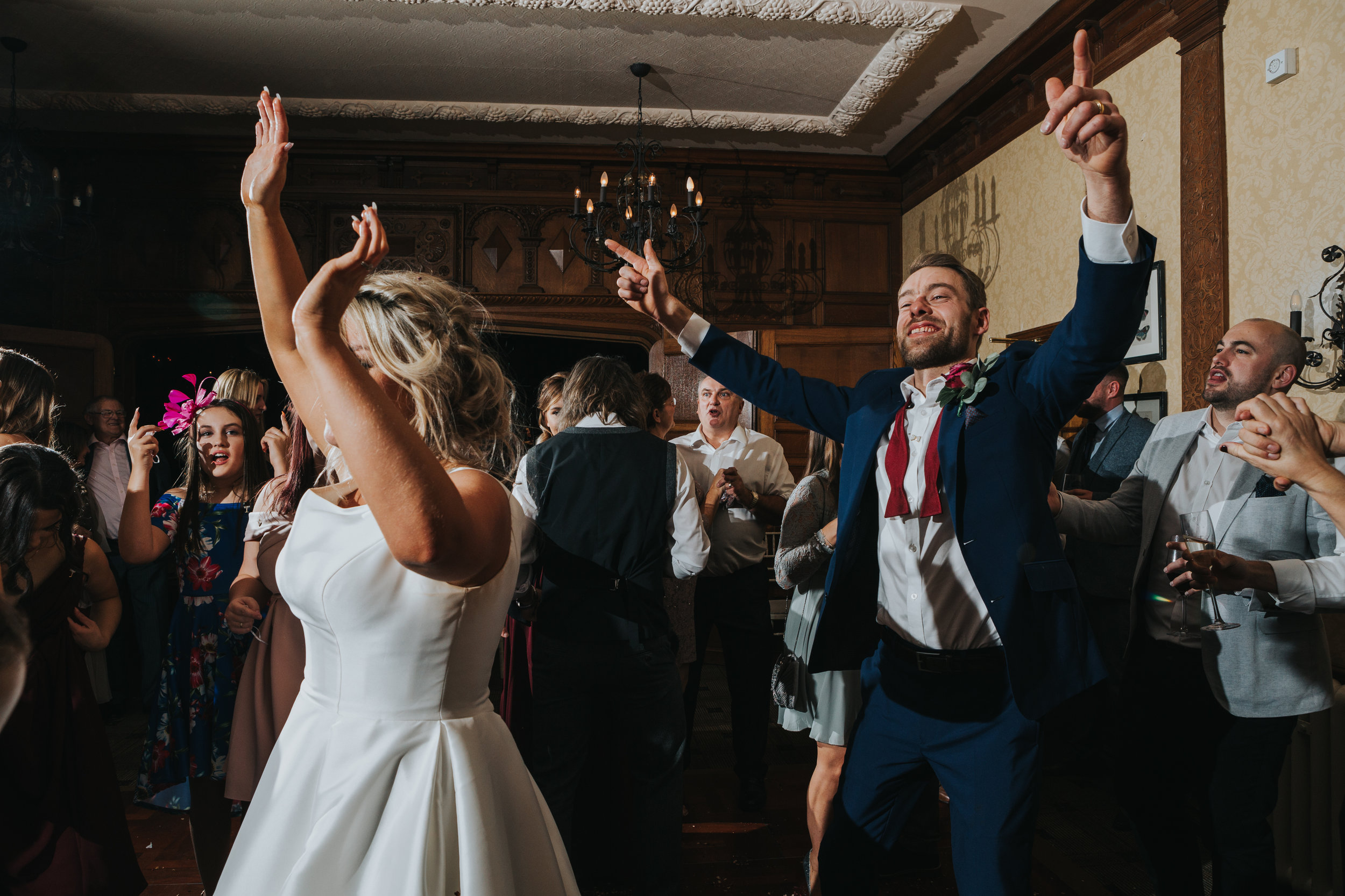 Groom stands nest to his new wife holding both hands in the air while grinning from ear to ear, surrounded by his friends and family. 