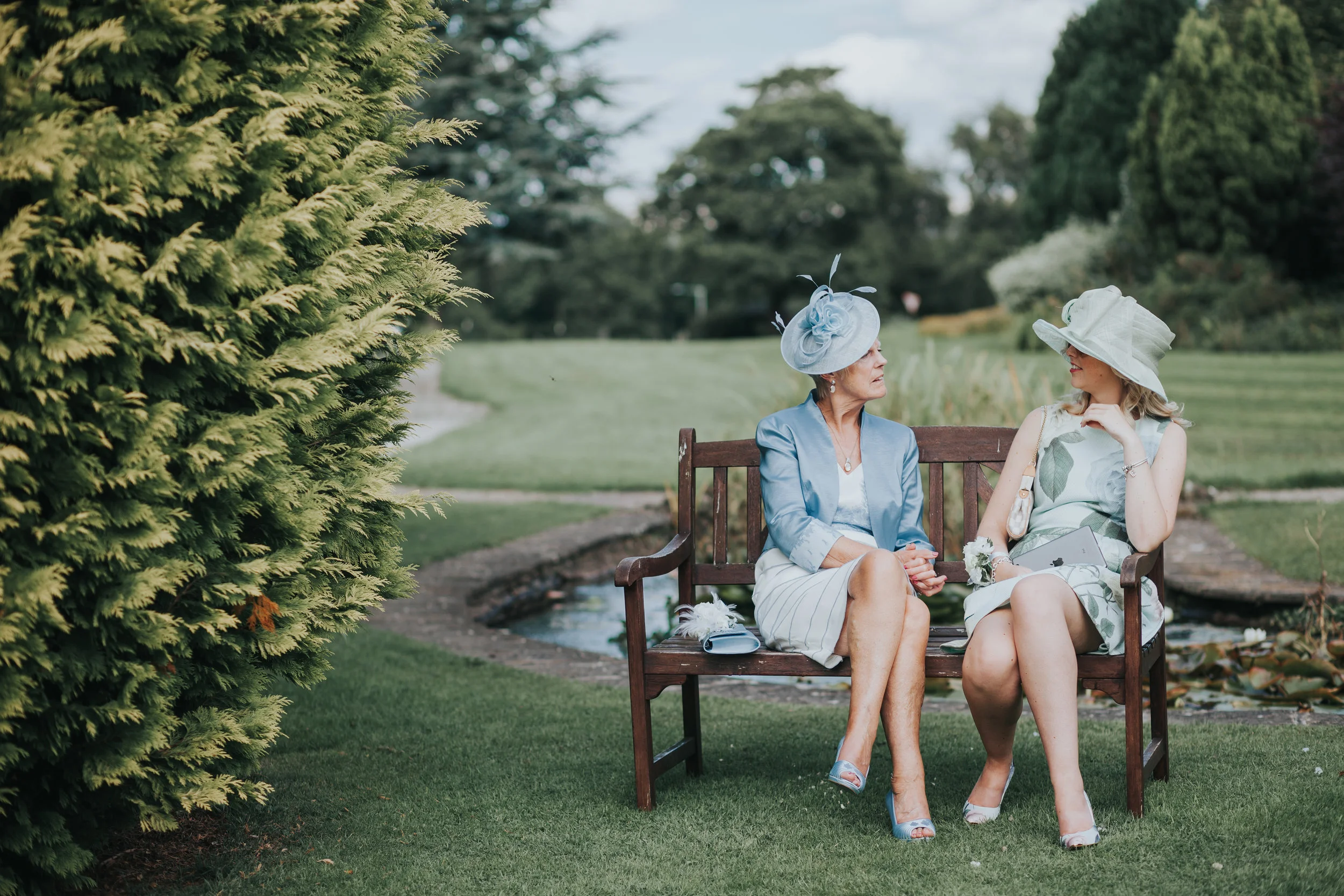 Wedding guests wearing pale green and blue outfits and hats sit together on the bench at Mytton Fold. 