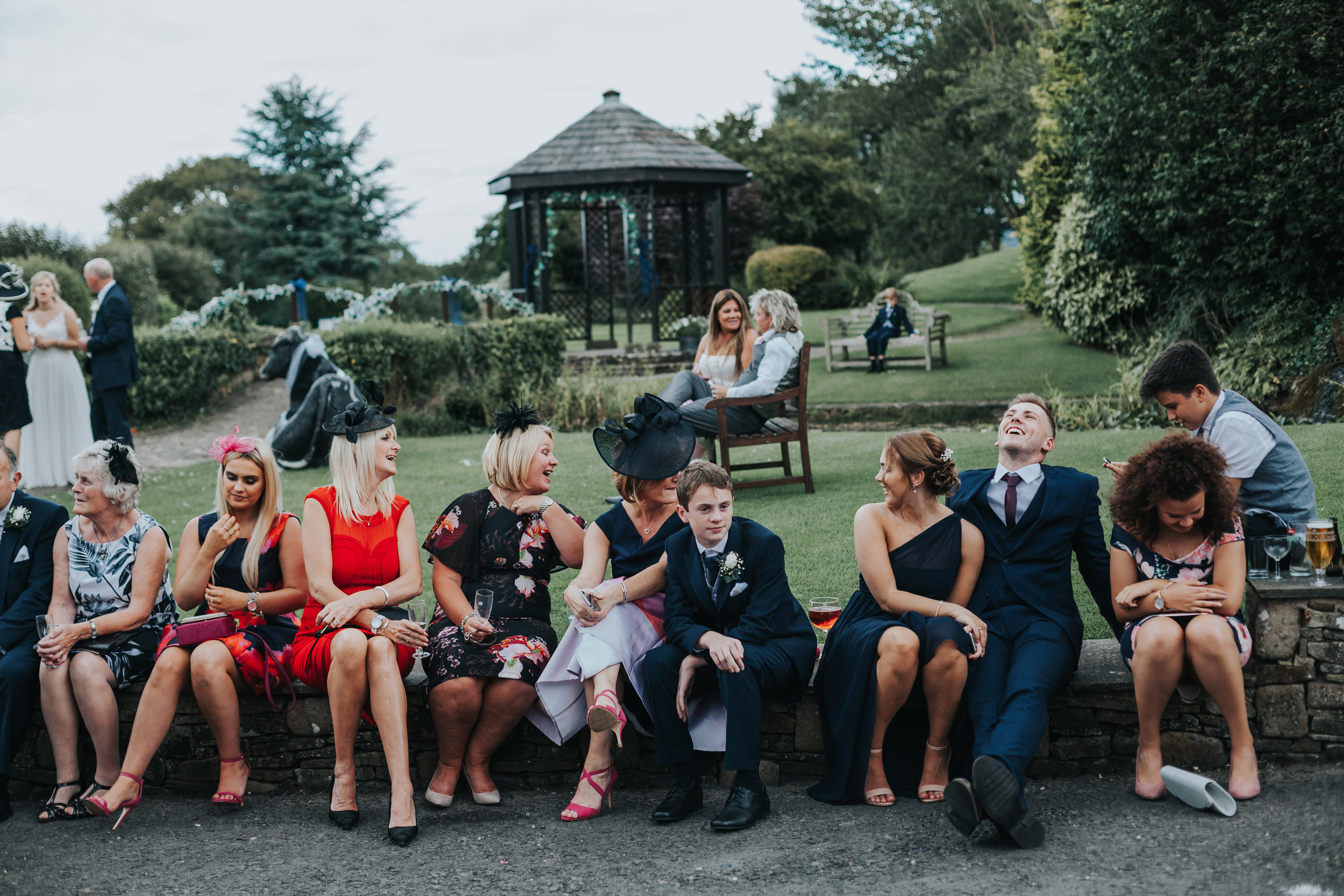 Wedding guests sit on a wall together laughing and drinking. 