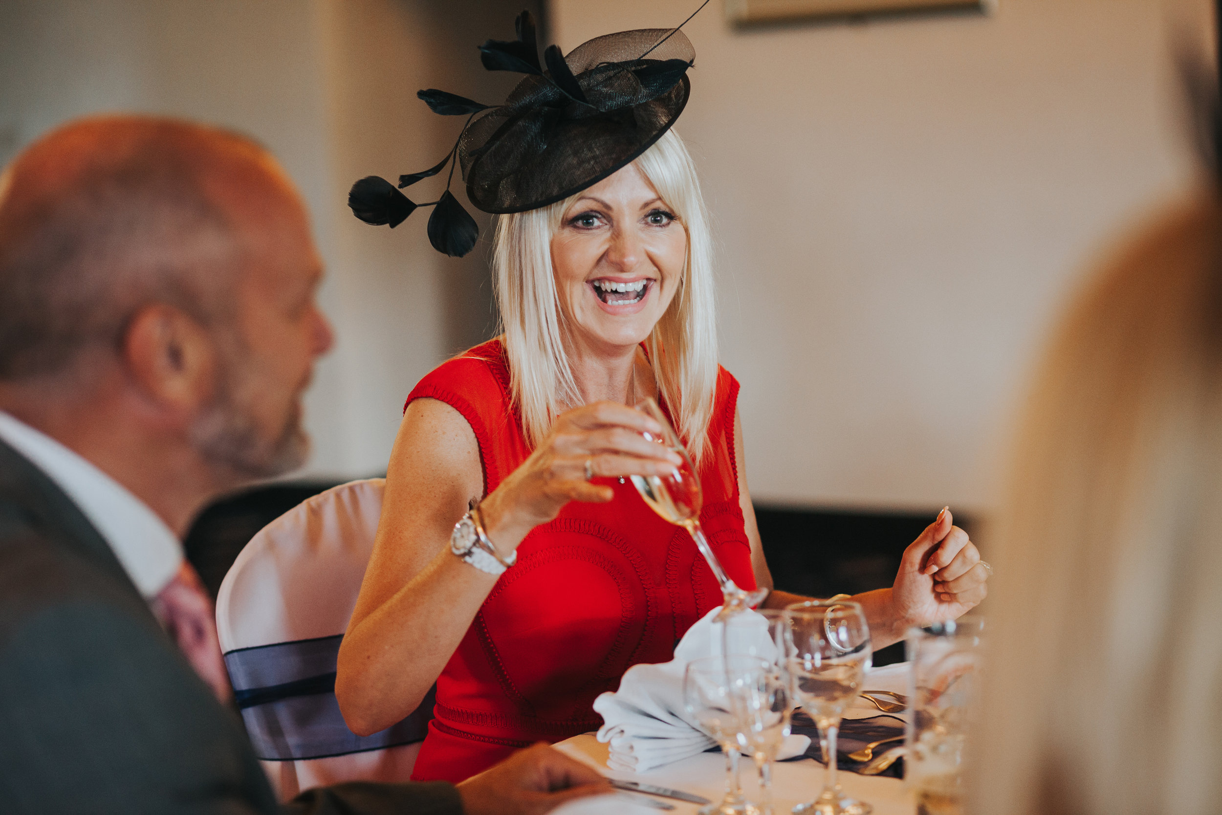 Wedding guest wears a red dress and black hat while laughing. 