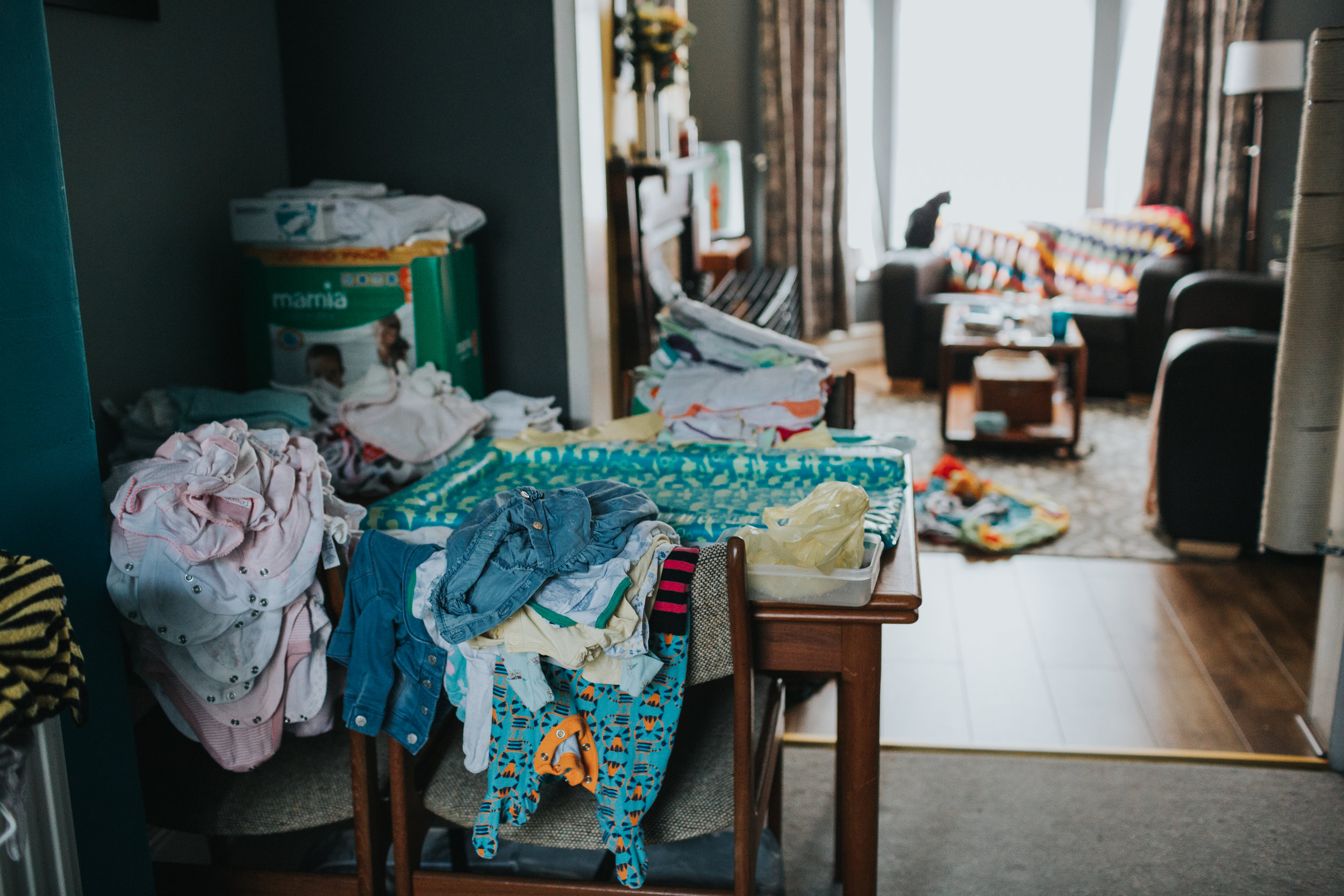 Baby clothes piled up on the dining table at the families house in Liverpool. In the back ground the family cat sits of the sofa looking out the window.