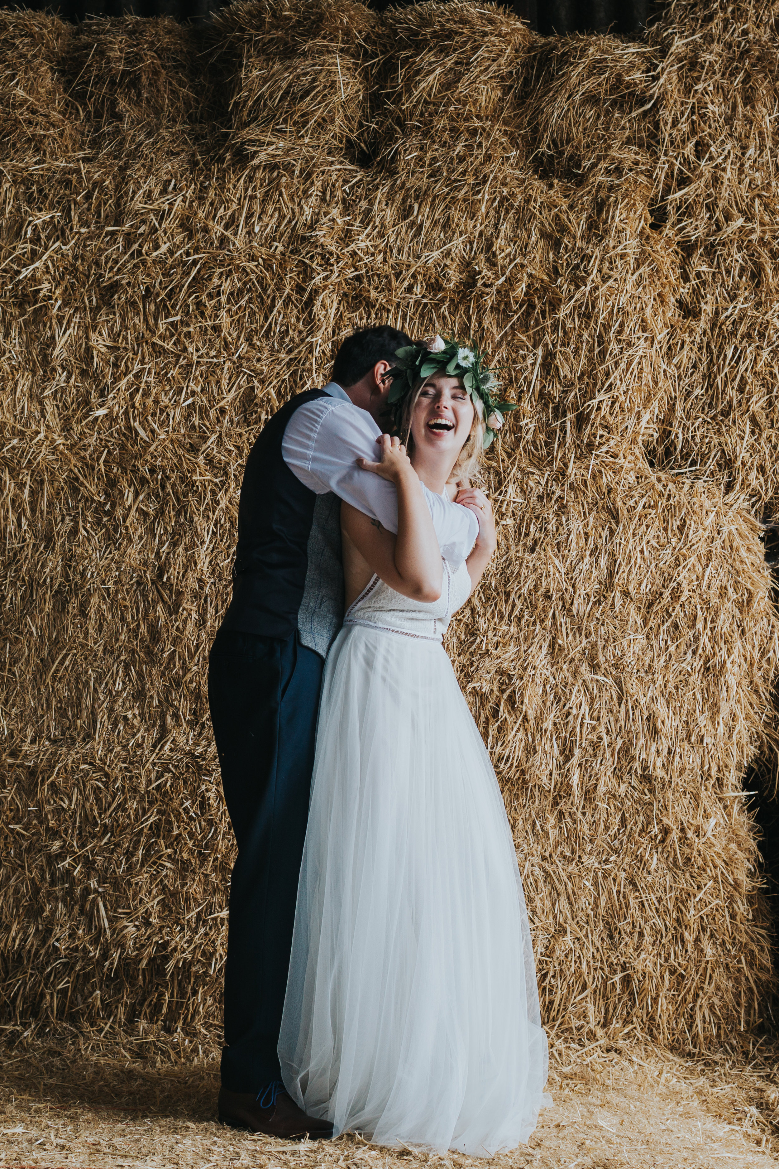 Bride and groom laugh together in front  of a load of hay stacks. 