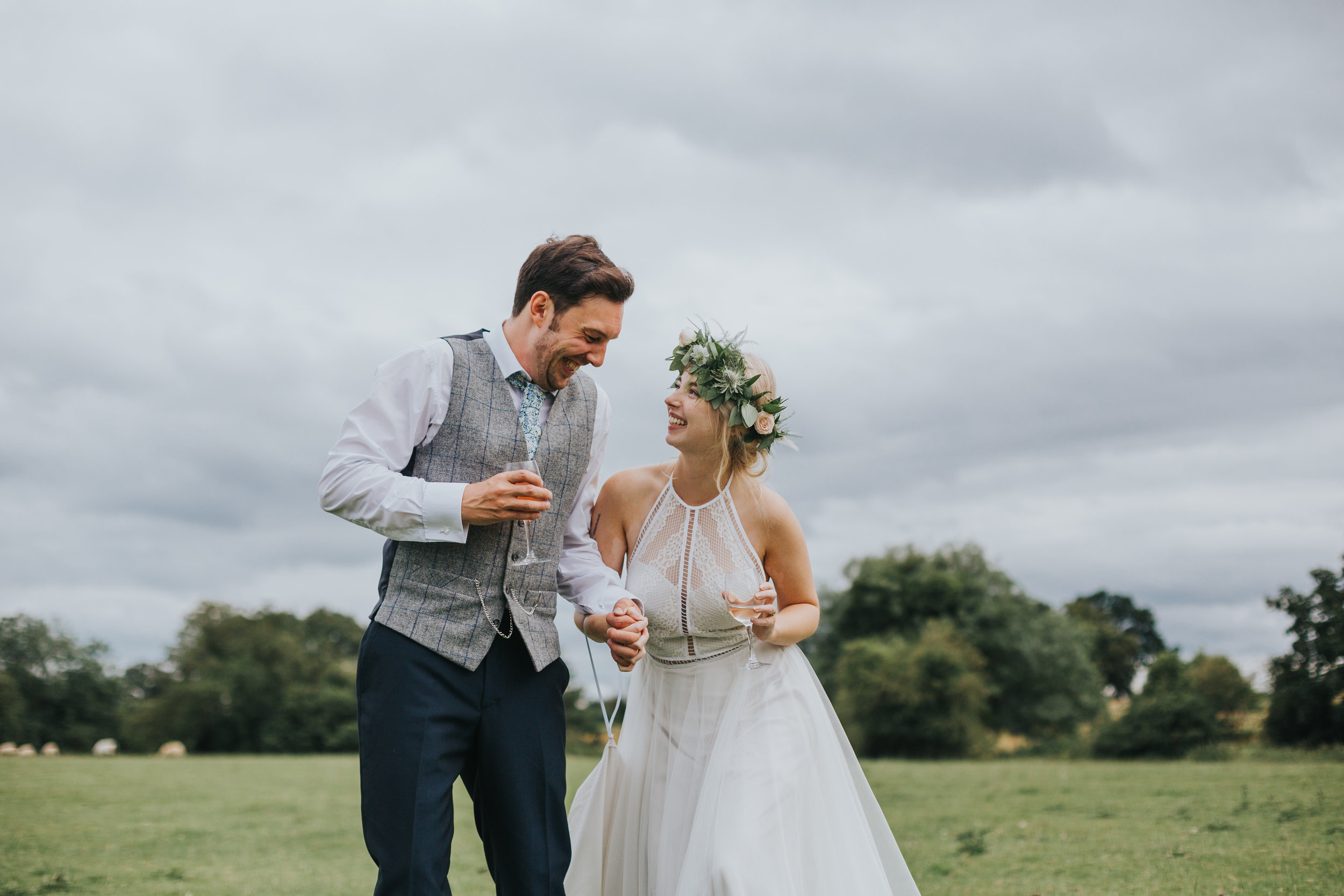 Bride and Groom sip champagne and laugh together walking through a field near Owen House Wedding Barn. 