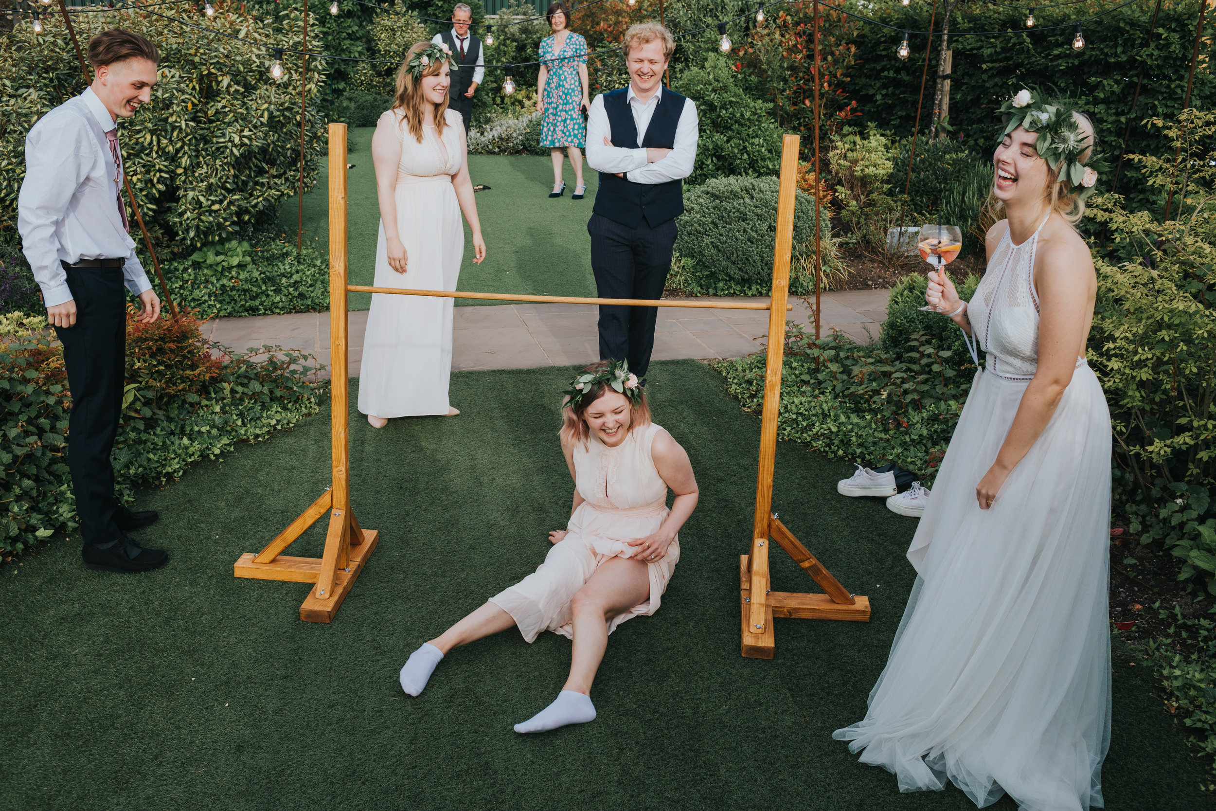 Bridesmaid falls on her bum while playing limbo. Bride is holding her drink and laughing.