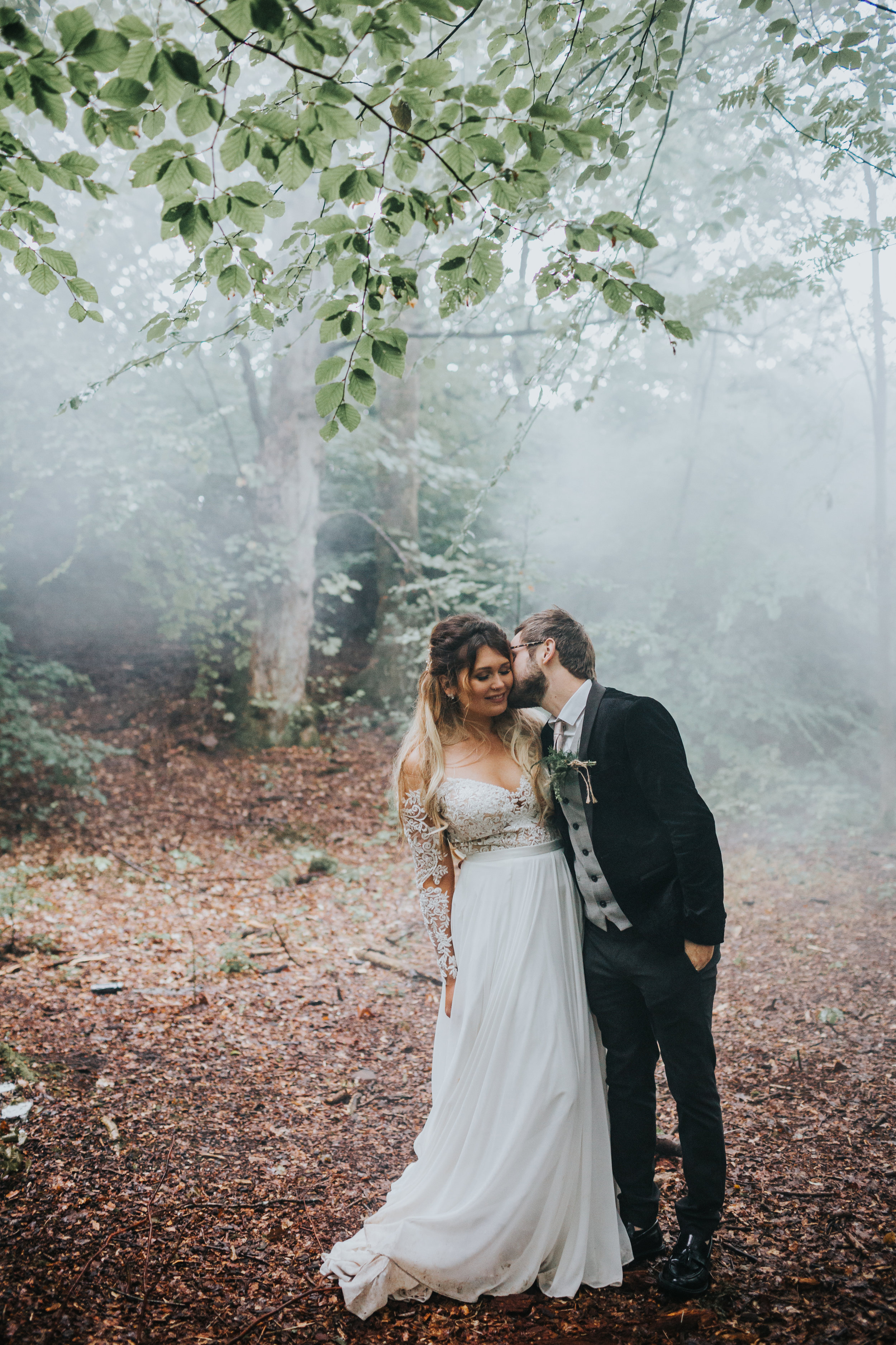 Groom kisses his bride in the woods with mist behind them. 