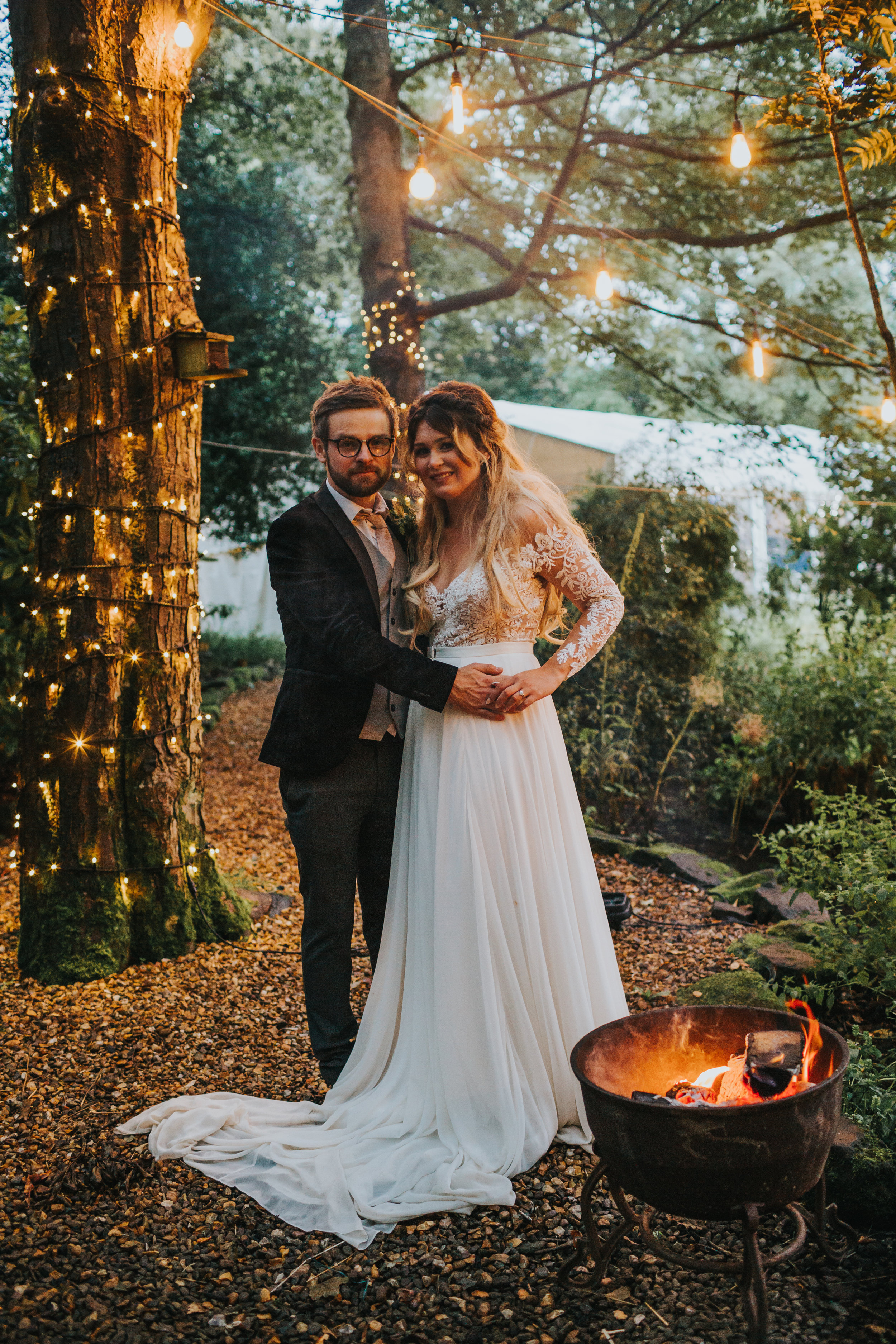 The bride and groom stand by the camp fire.