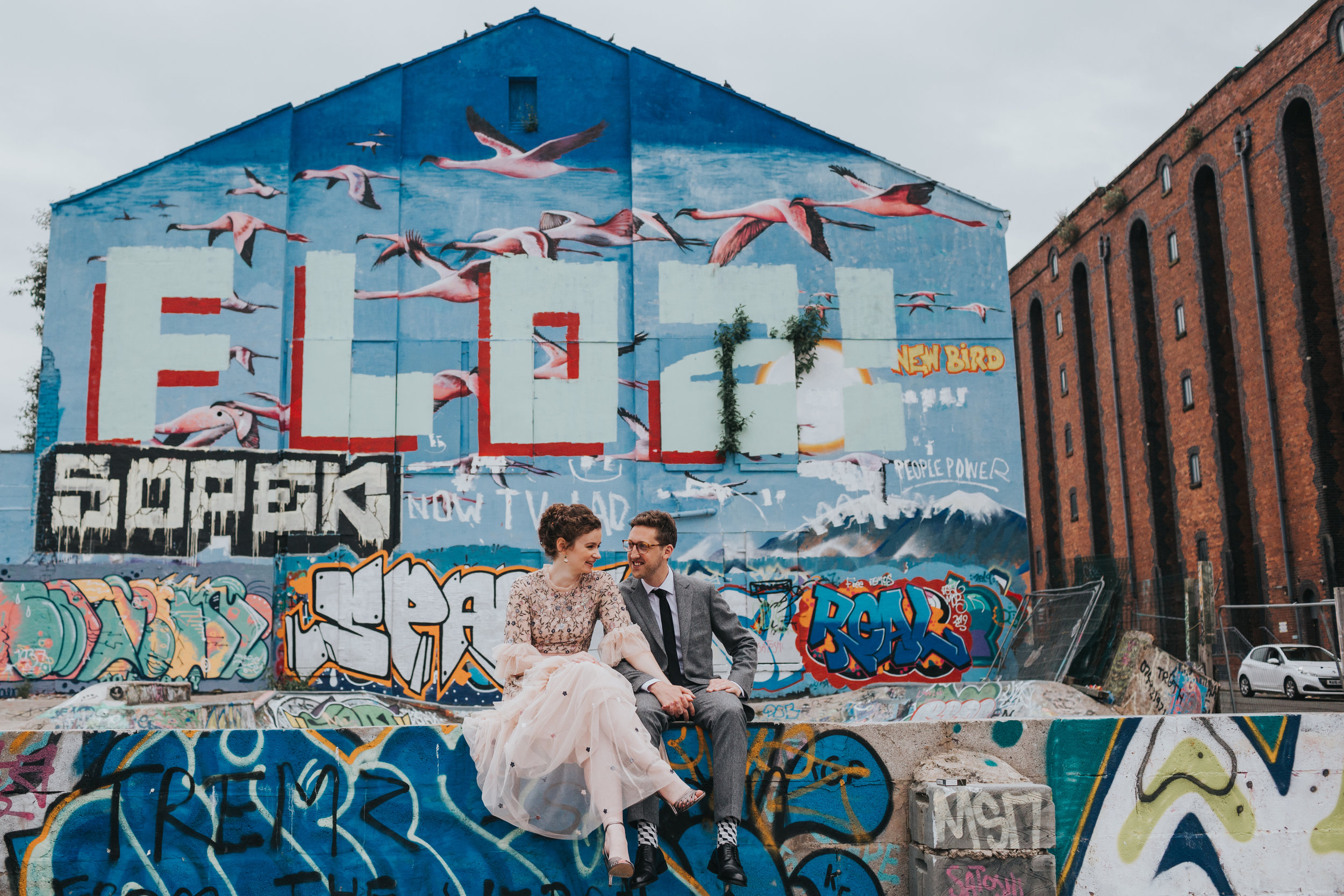 Bride and Groom sit in front of a blue and pink mural of a flock of flying flamingos. 