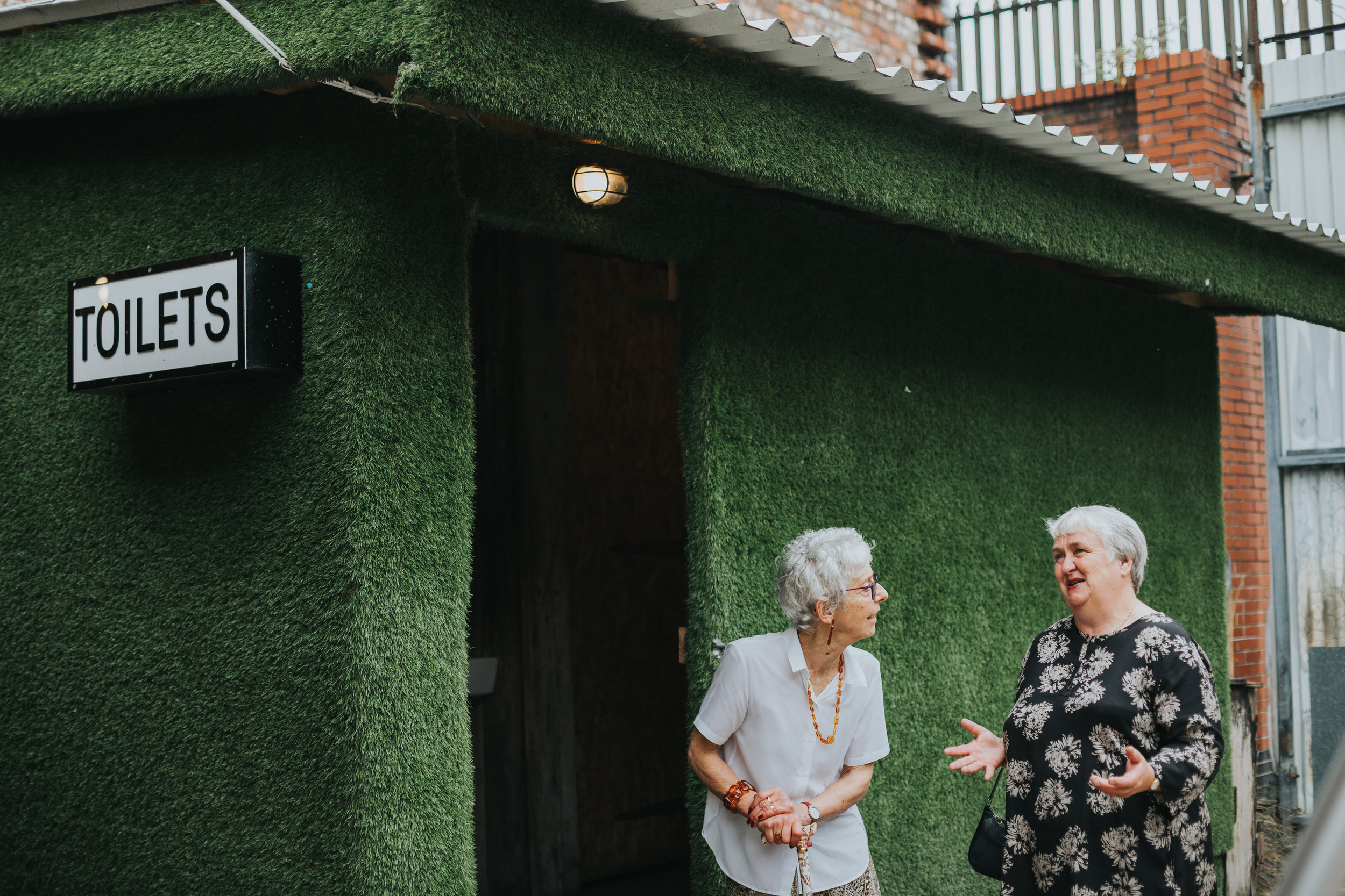 Two older female wedding guests chat outside the toilets. 