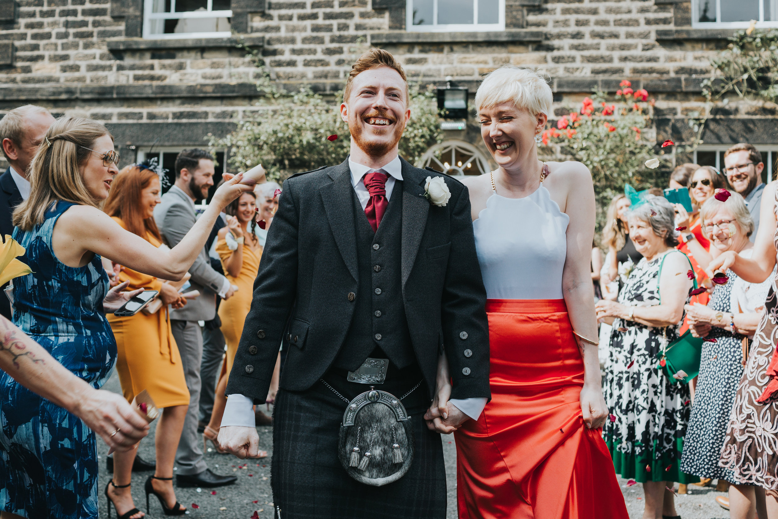 Bride and groom laugh as red petal confetti is thrown at them. 