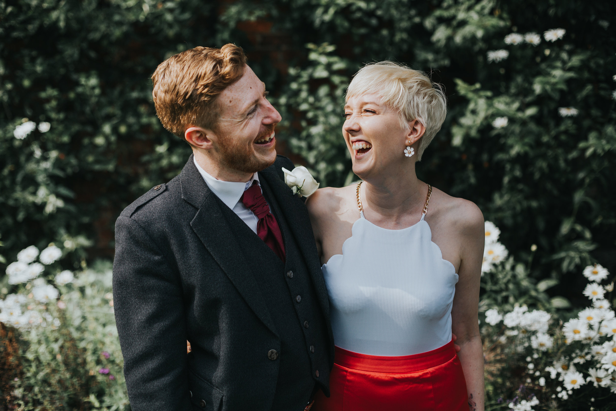 Bride and groom laughing in front of flowers. 