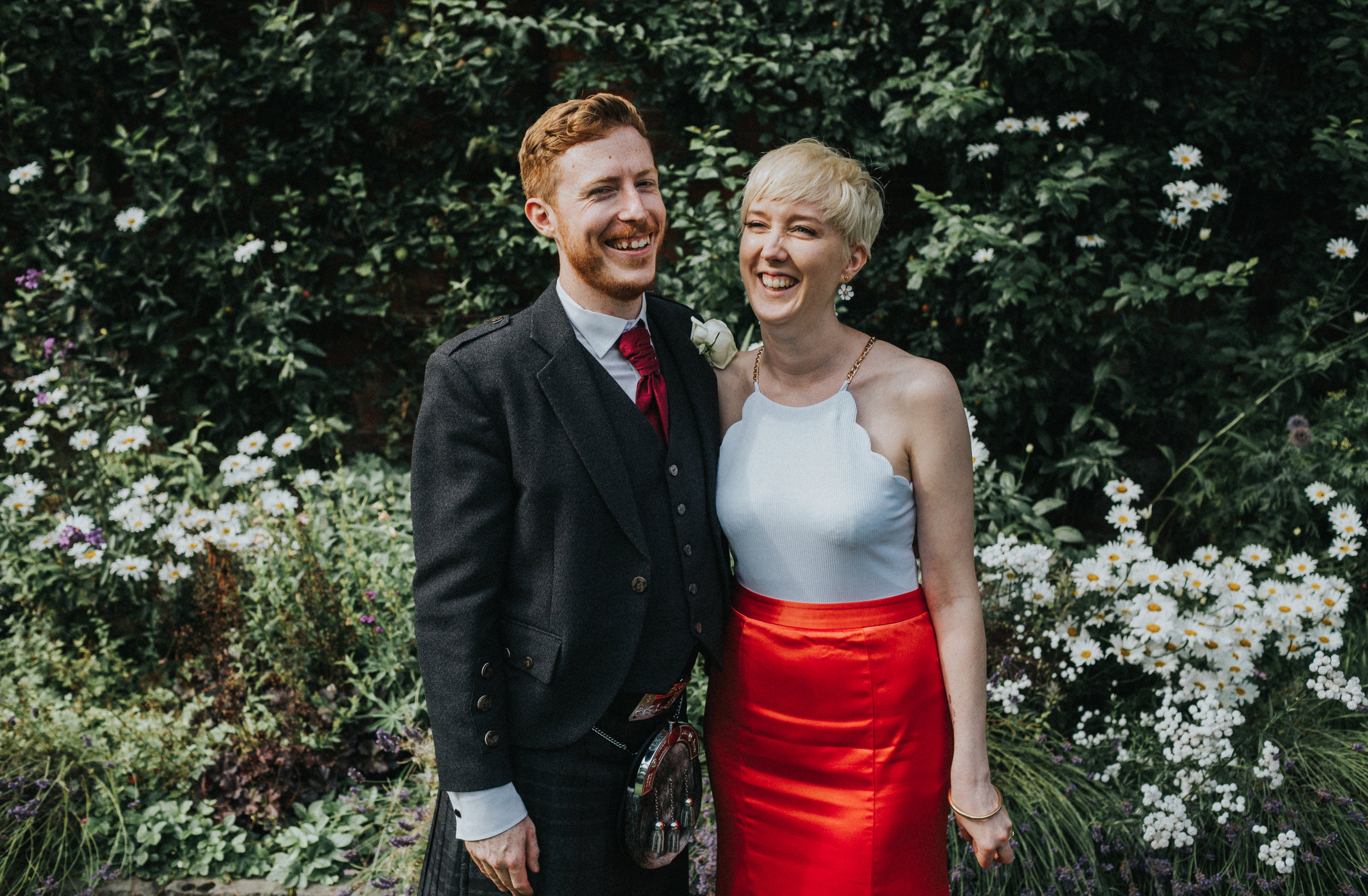 Bride and groom stand together in front of a row of daisies, bride wears daisy ear rings.
