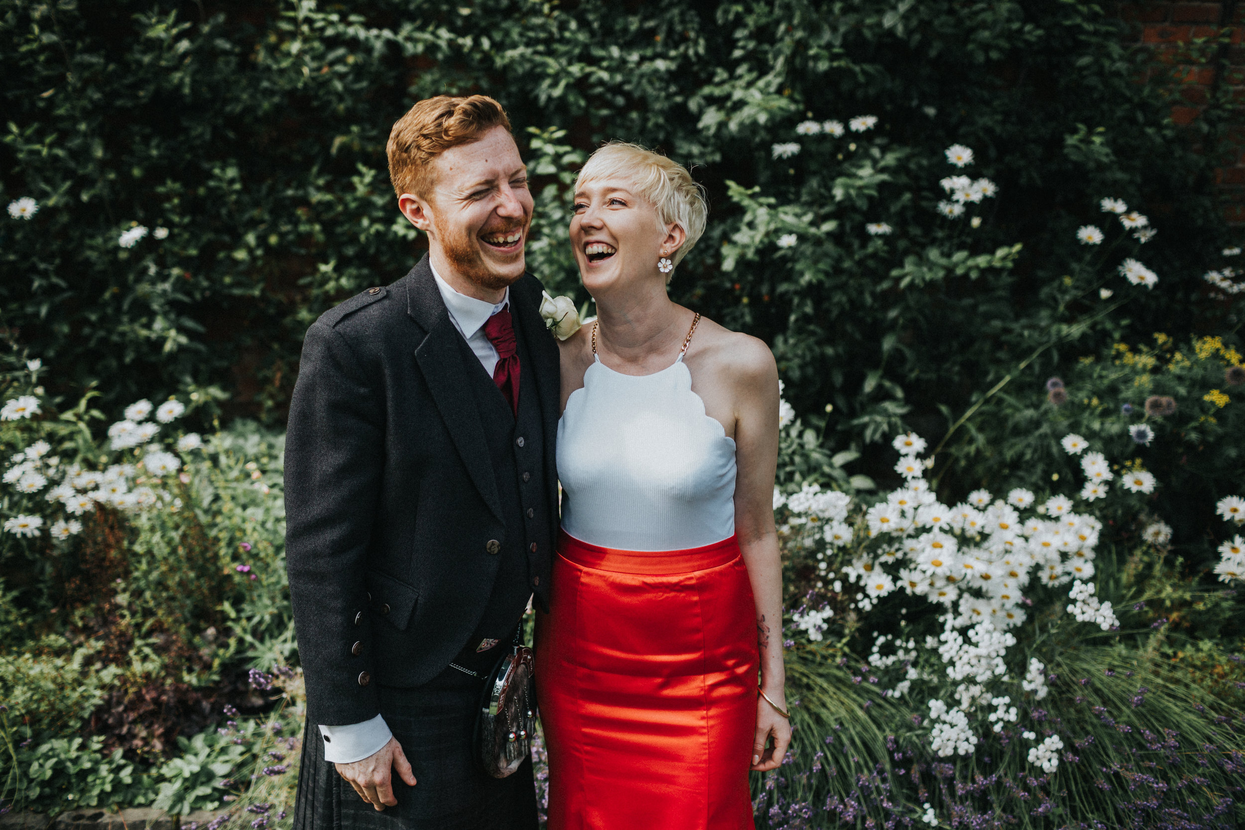 Untraditional bride and groom stand laughing on their chilled out wedding celebration. 