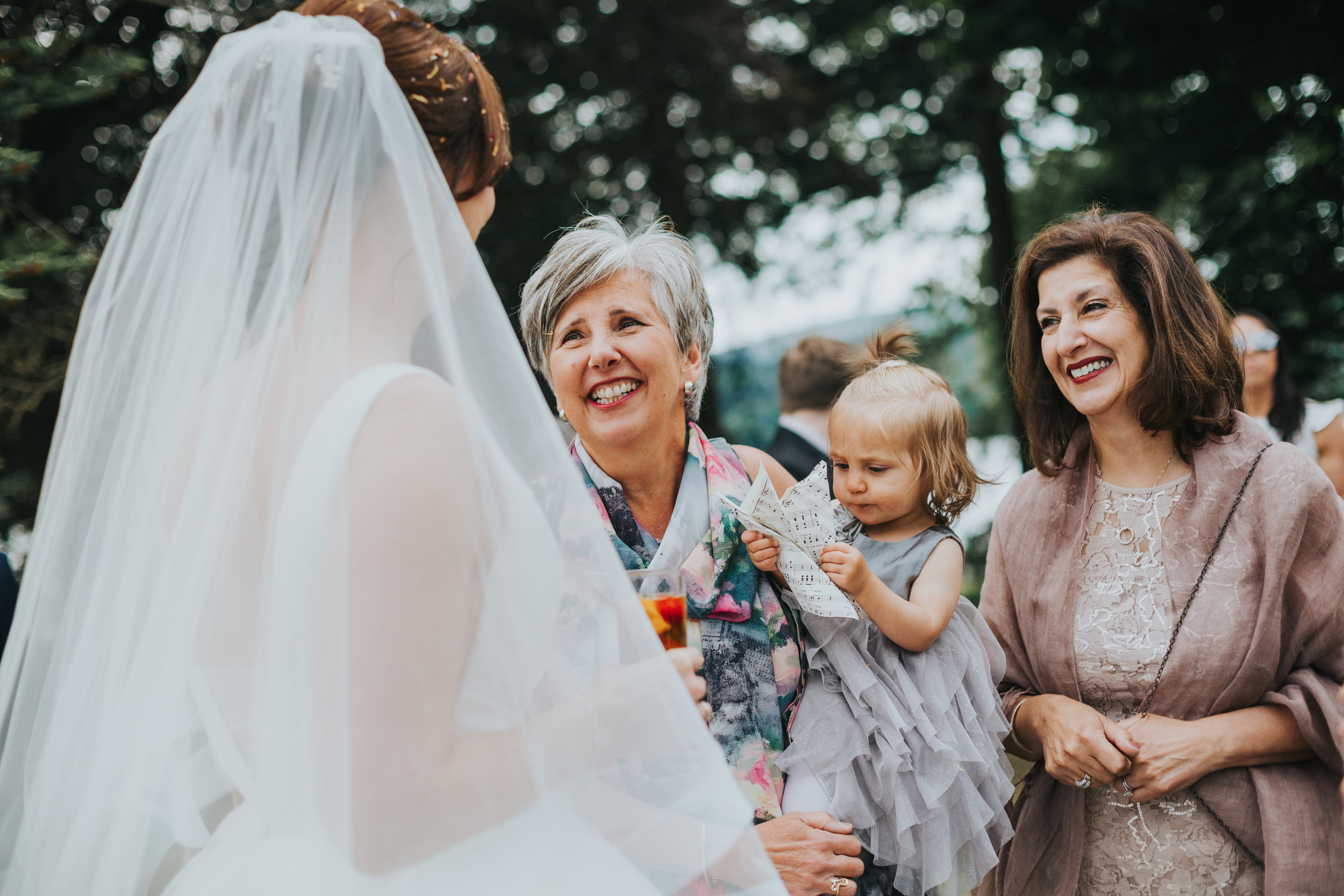 Bride greets smiling wedding guests holding a baby. 