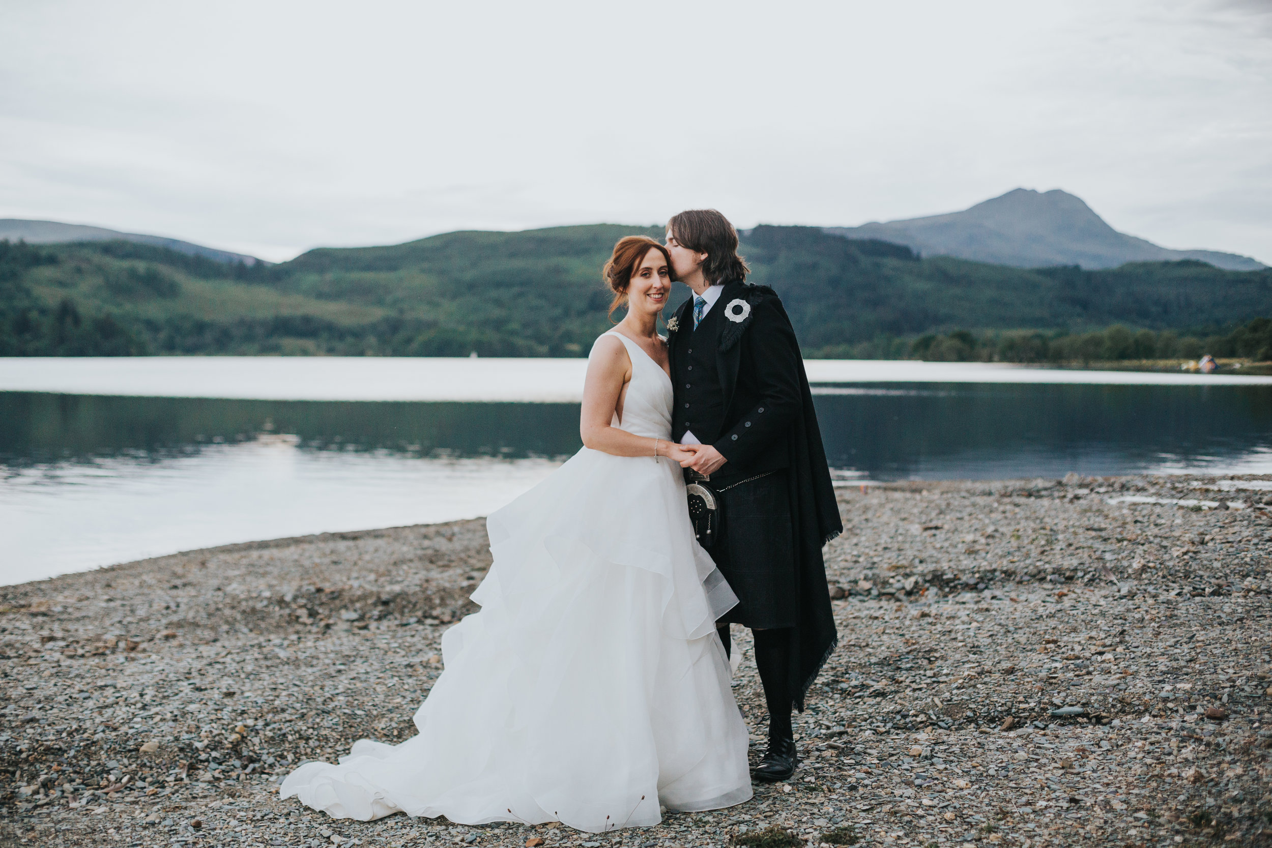 Bride and groom stand on beach of Loch Ard.