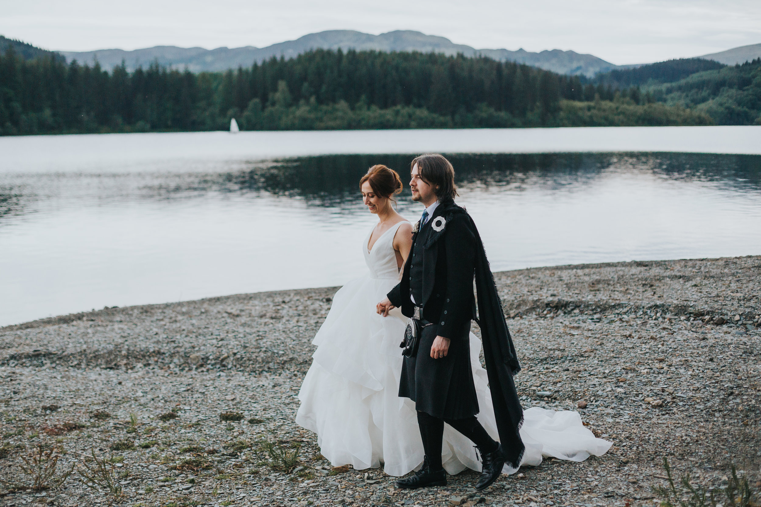 Bride and Groom walk along bank of Loch Ard. 