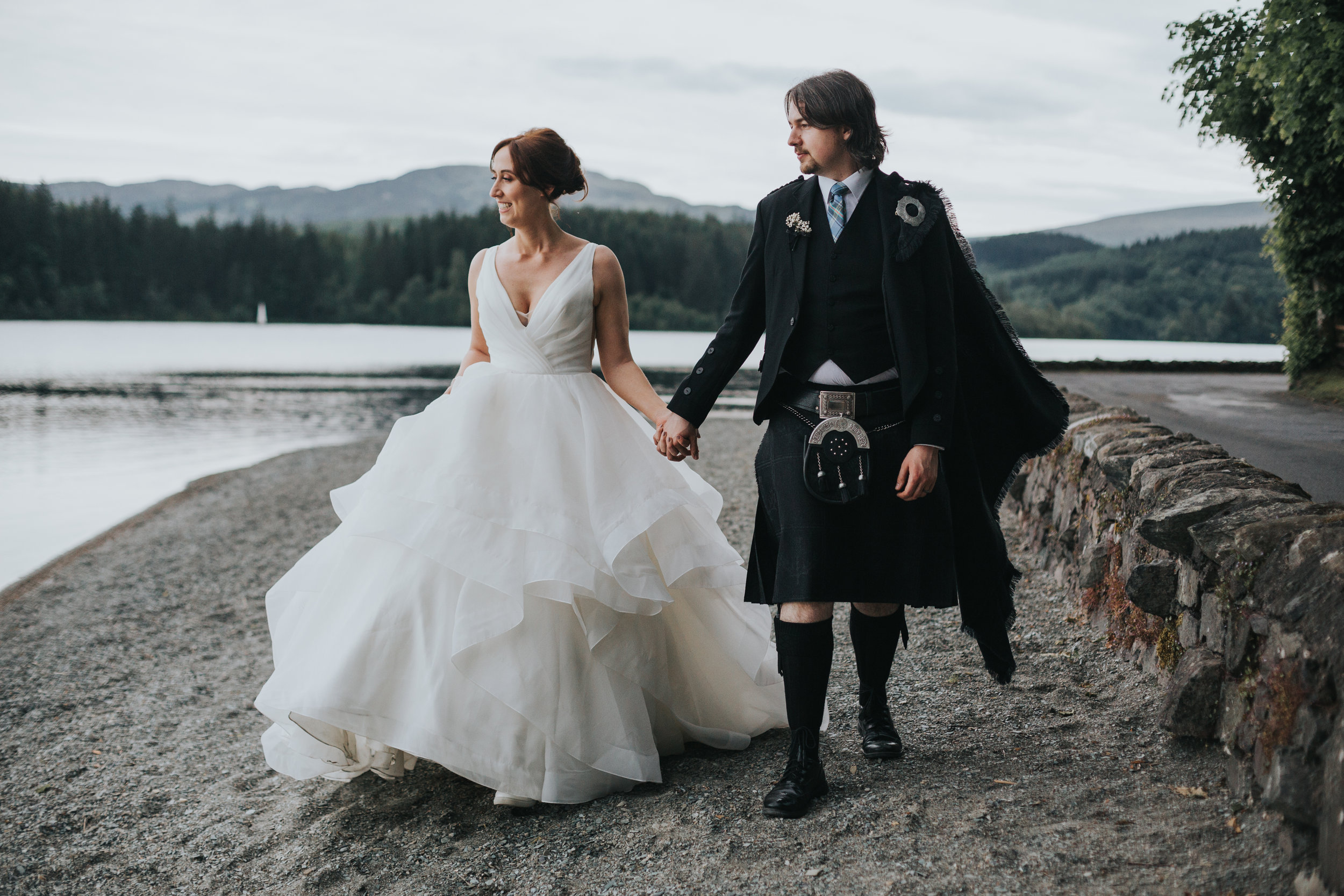Bride and Kilted Groom walk together along the pebbled beach at Loch Ard, Scotland. 