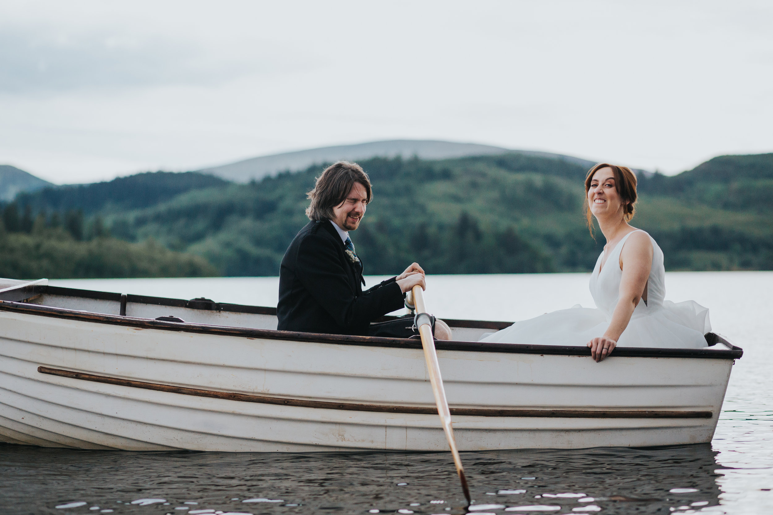 Bride pulls a funny face while groom laugh in rowing boat on Loch Ard. 