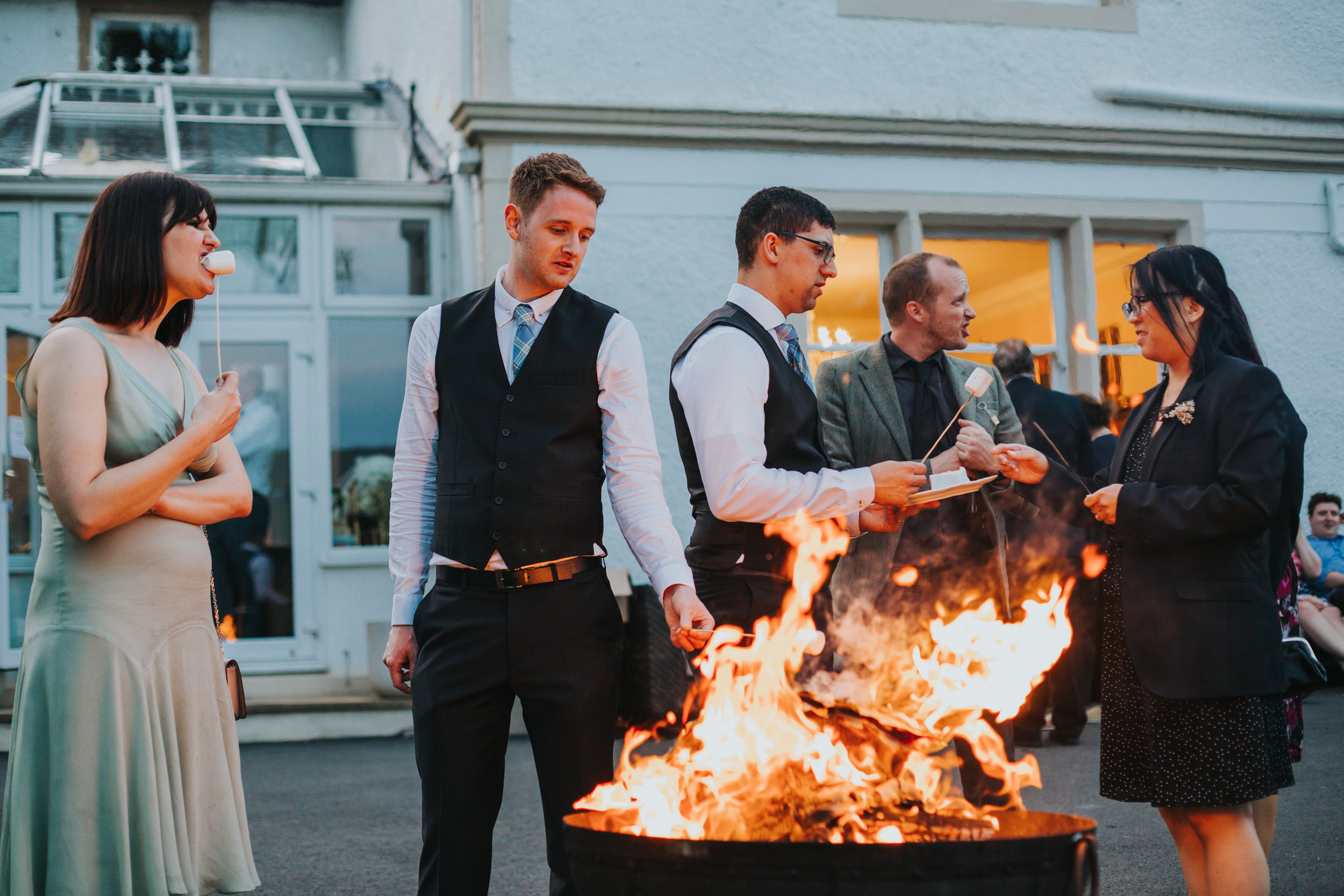 Wedding guest munches on marsh mellow while other wedding guests toast theirs of fire .