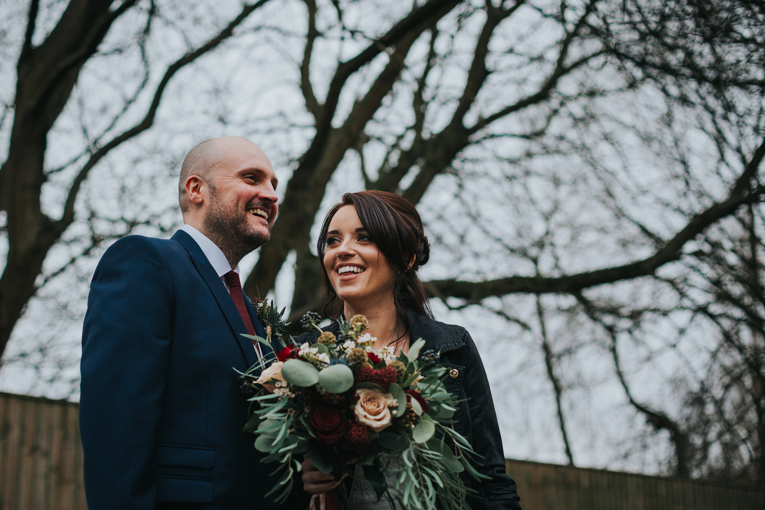 Bride and groom smiling with wintery sky and bare branches behind them. 