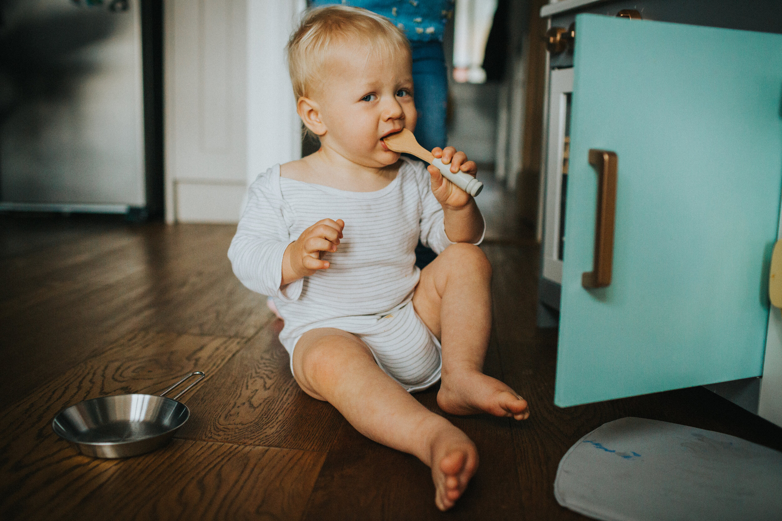 Little boy bites a wooden spoon while peering at the camera. 
