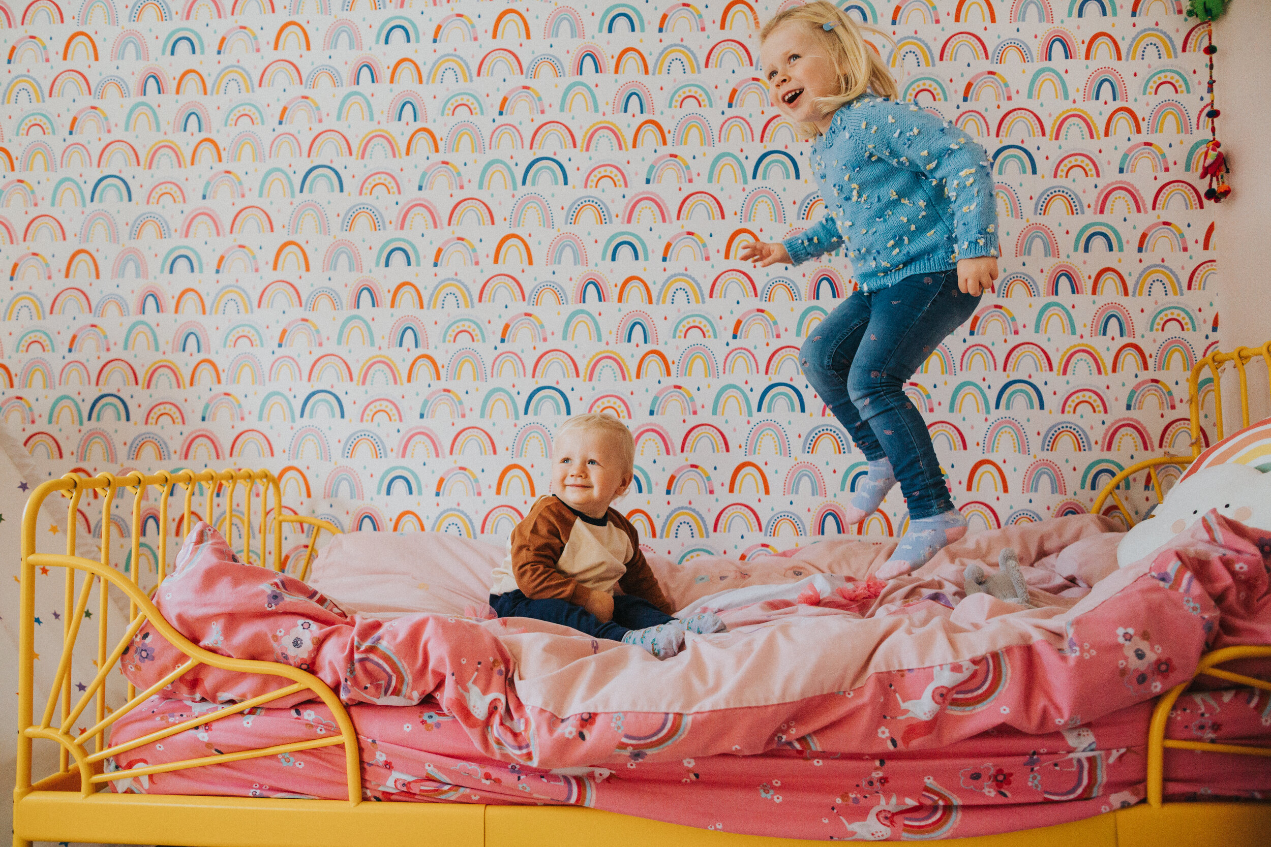 Little girl jumps on her bed with her little brother in brightly coloured rainbow wall papered room.