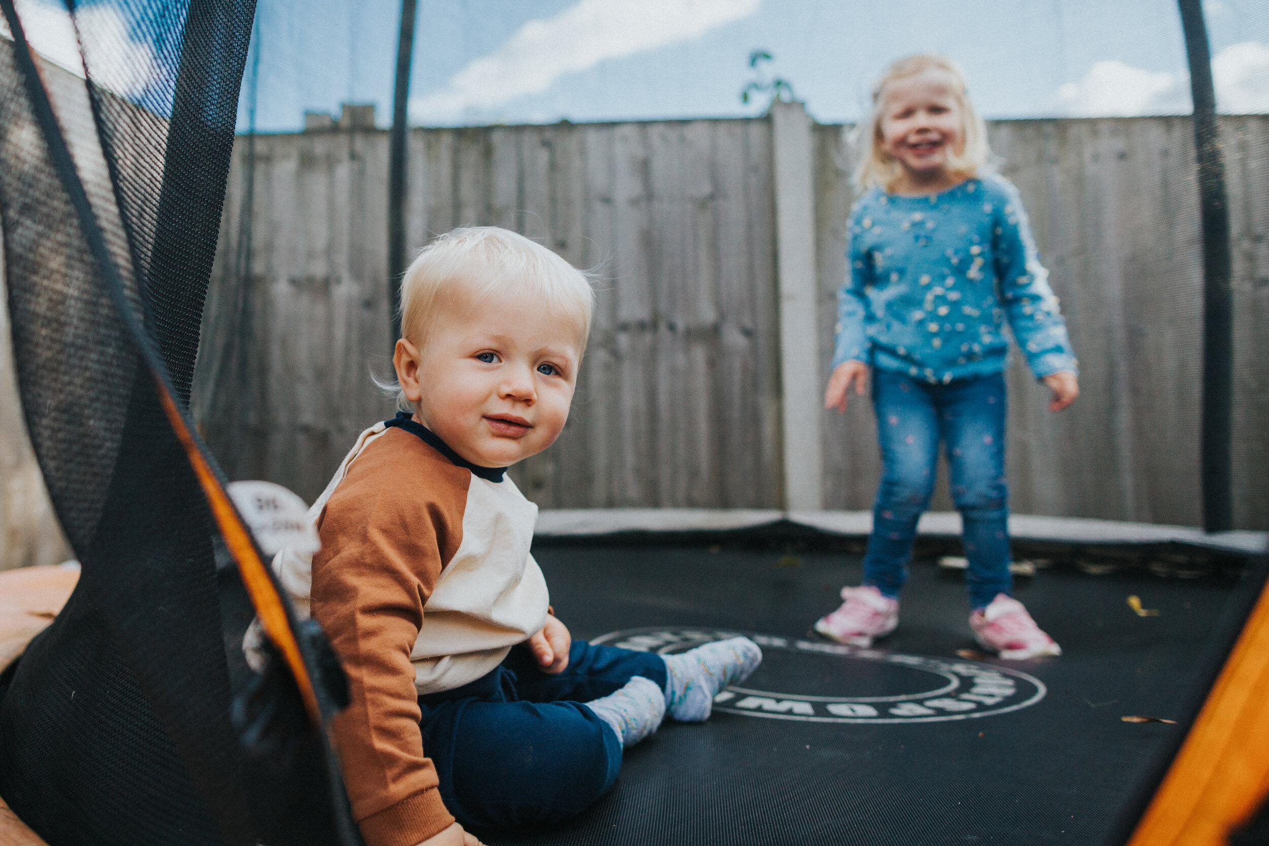 Little boy sits on trampoline as his sister plays. 