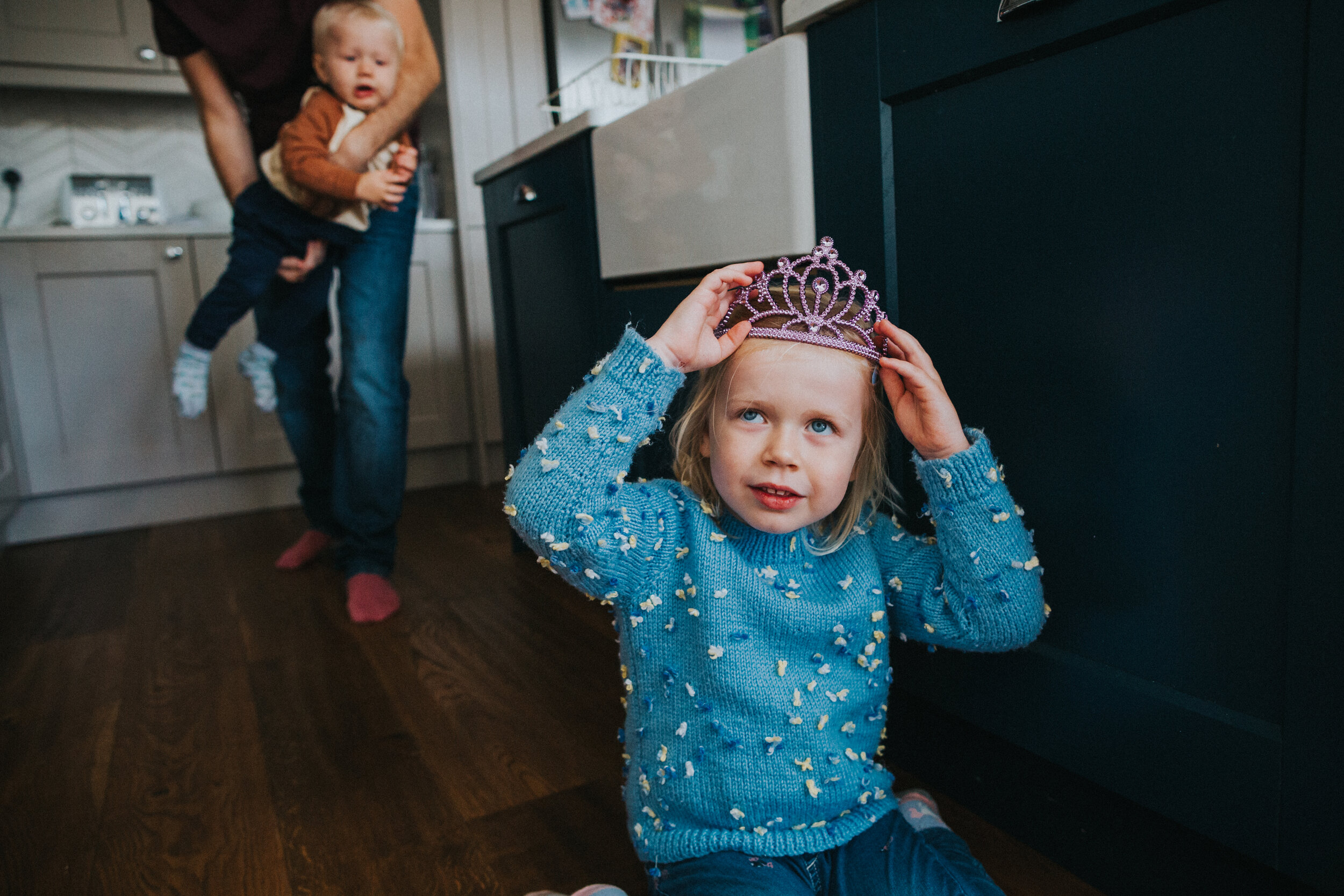 Little girl puts on crown as Dad approaches with her brother. 