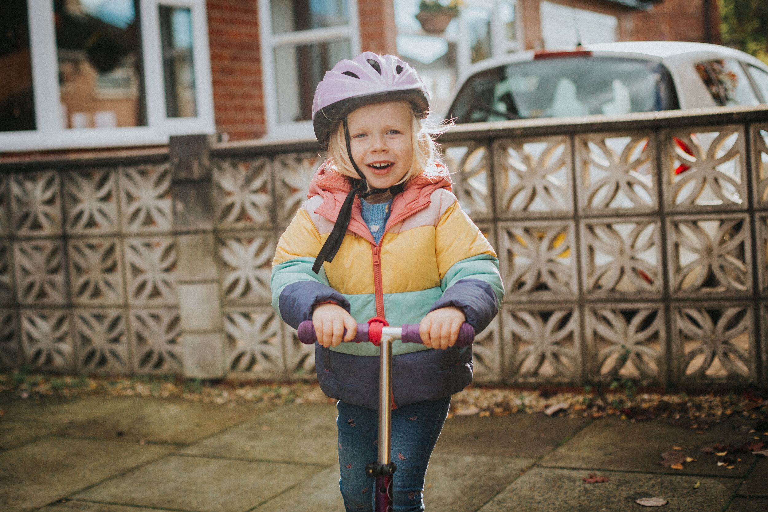 Little girl rides scooter with her purple crash helmet on and colourful coat. 