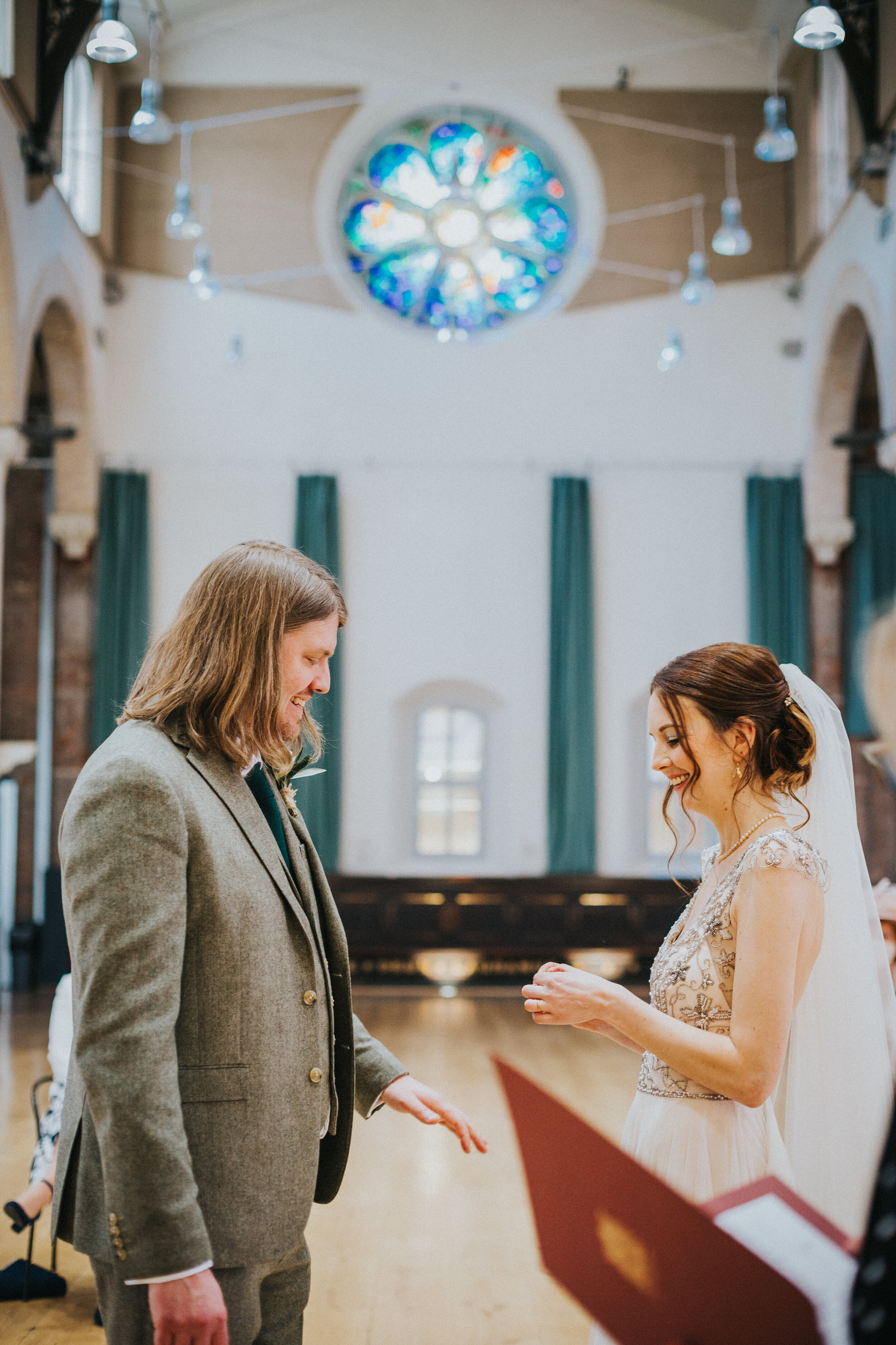 Couple exchange rings with a stained glass window behind them at Halle St Peters Manchester