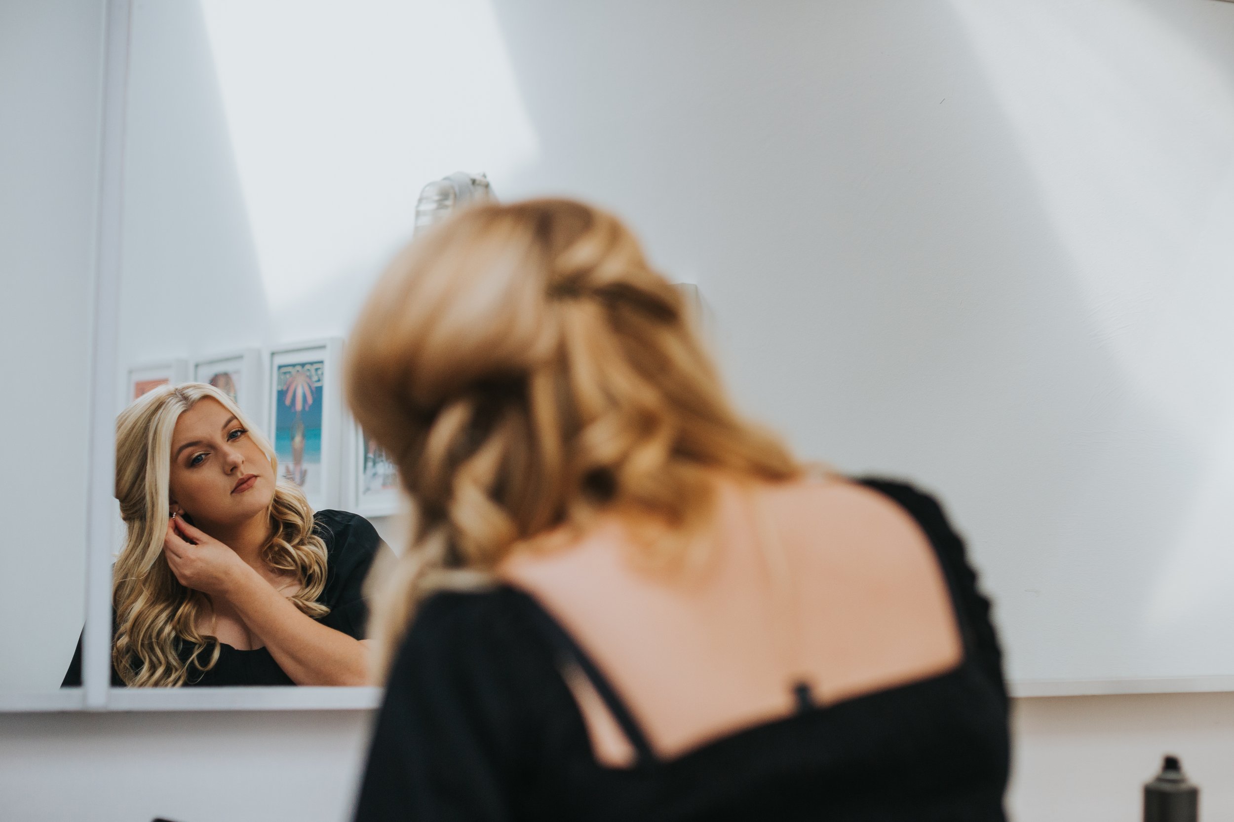 Blonde bridesmaid puts on earings in the mirror. 