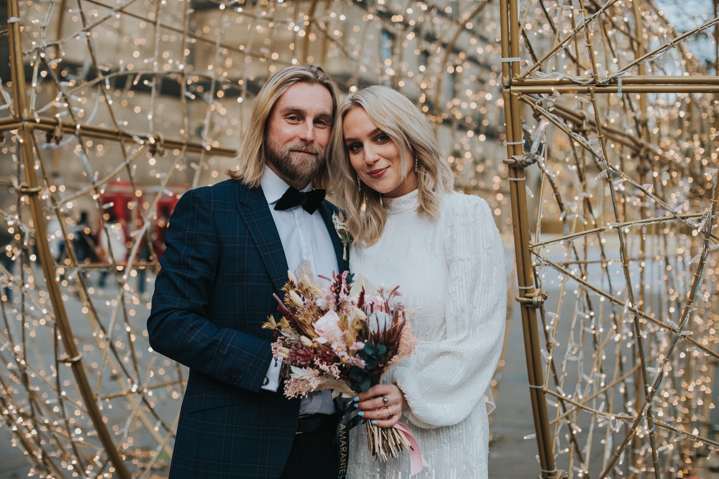 Bride and groom stand in an archway of golden Christmas lights. 