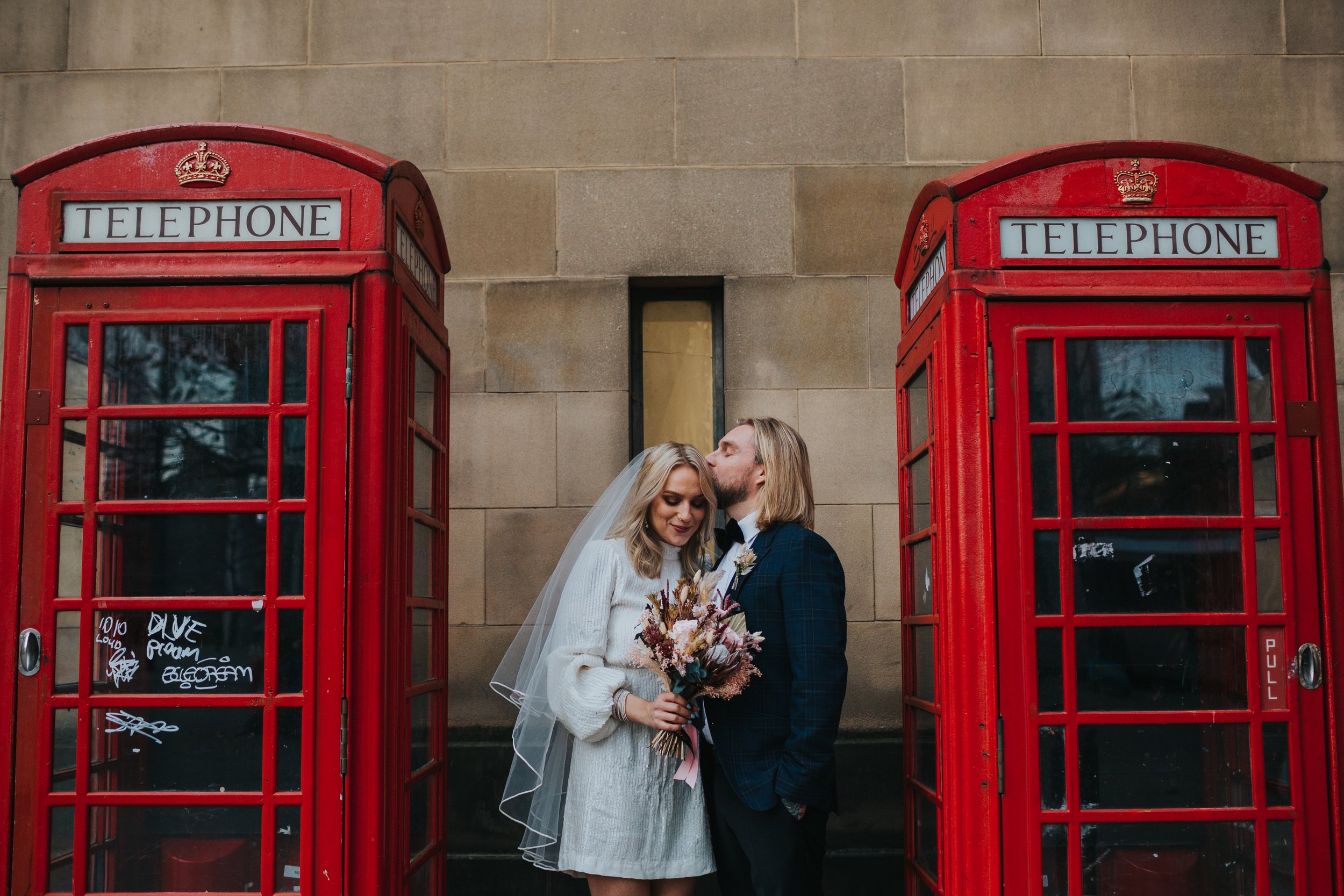 Groom gives his wife a kiss in between some red phone boxes in Manchester