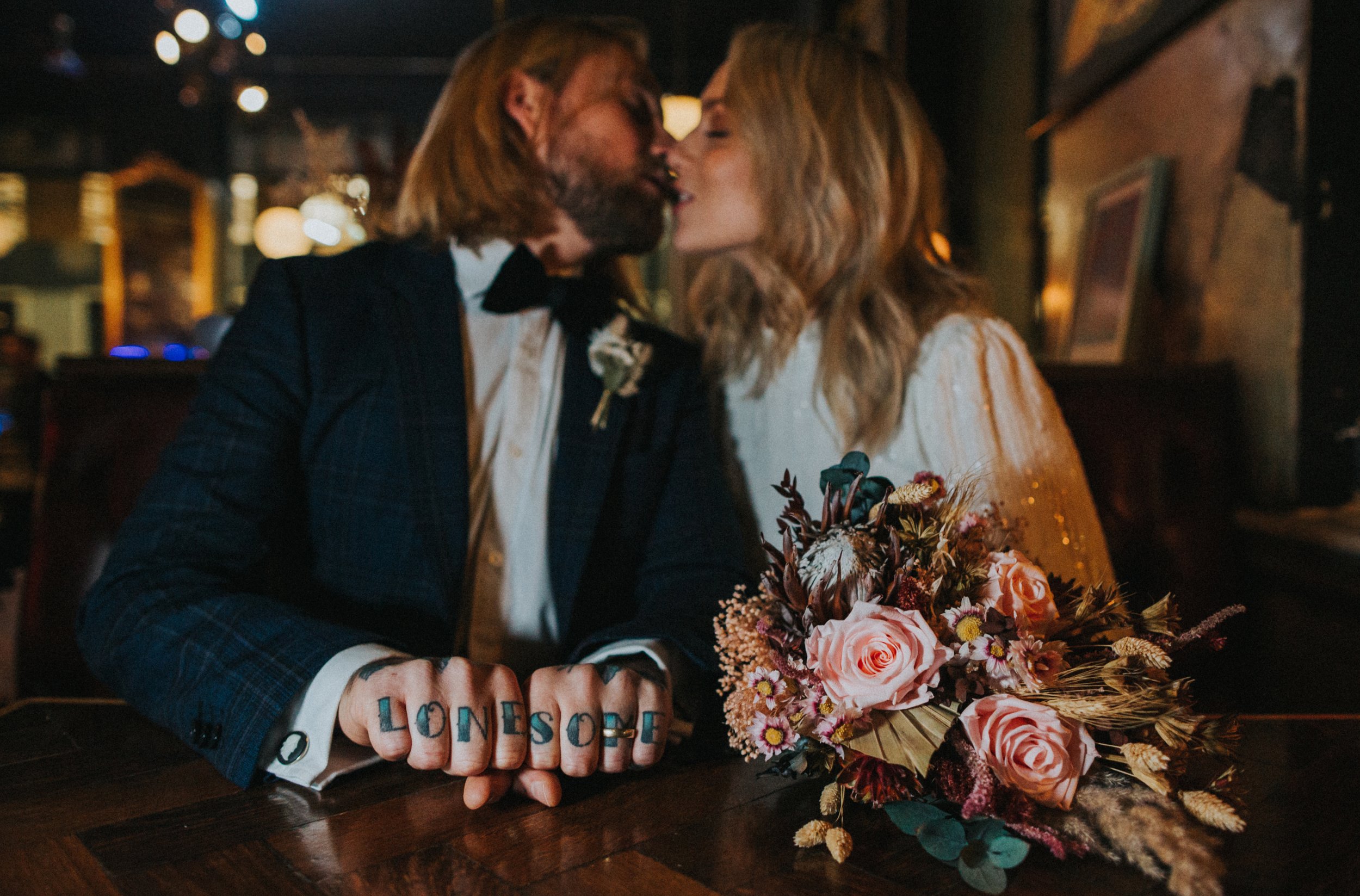 Groom and his "lonesome" knuckle tattoos and brides flowers together as the couple kiss in the back ground. 