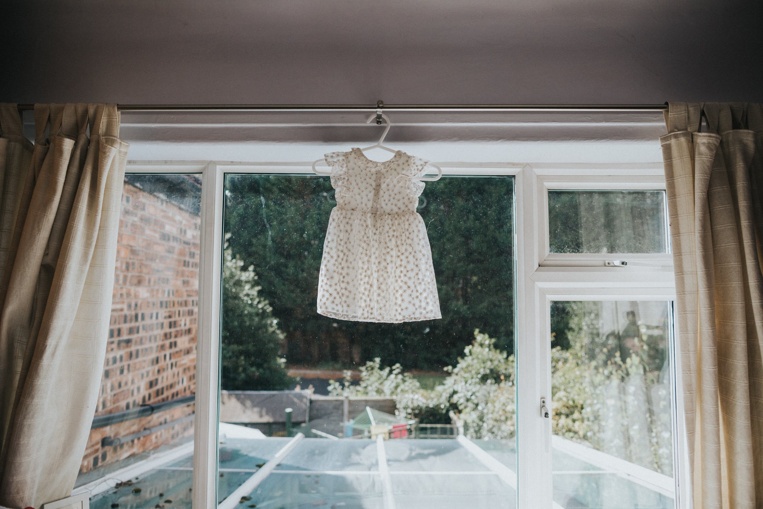 Brides daughters tiny white floral dress haging up in the brides childhood bedroom window.