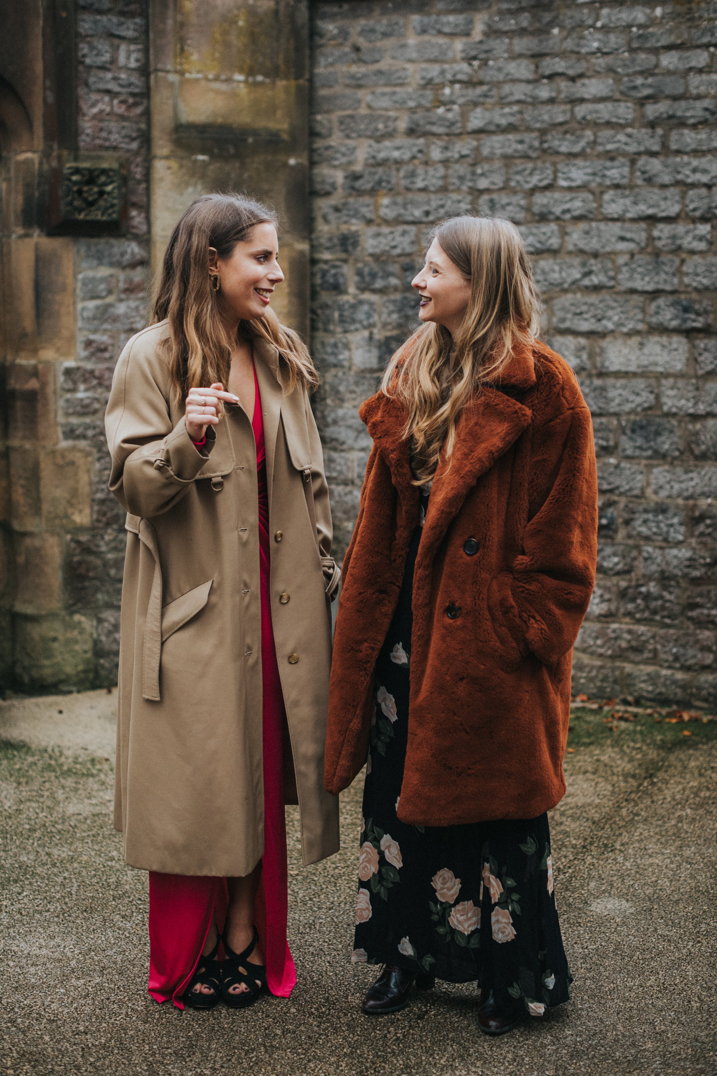 Two female wedding guests dressed in fancy winter coats chat outside the wedding venue. 