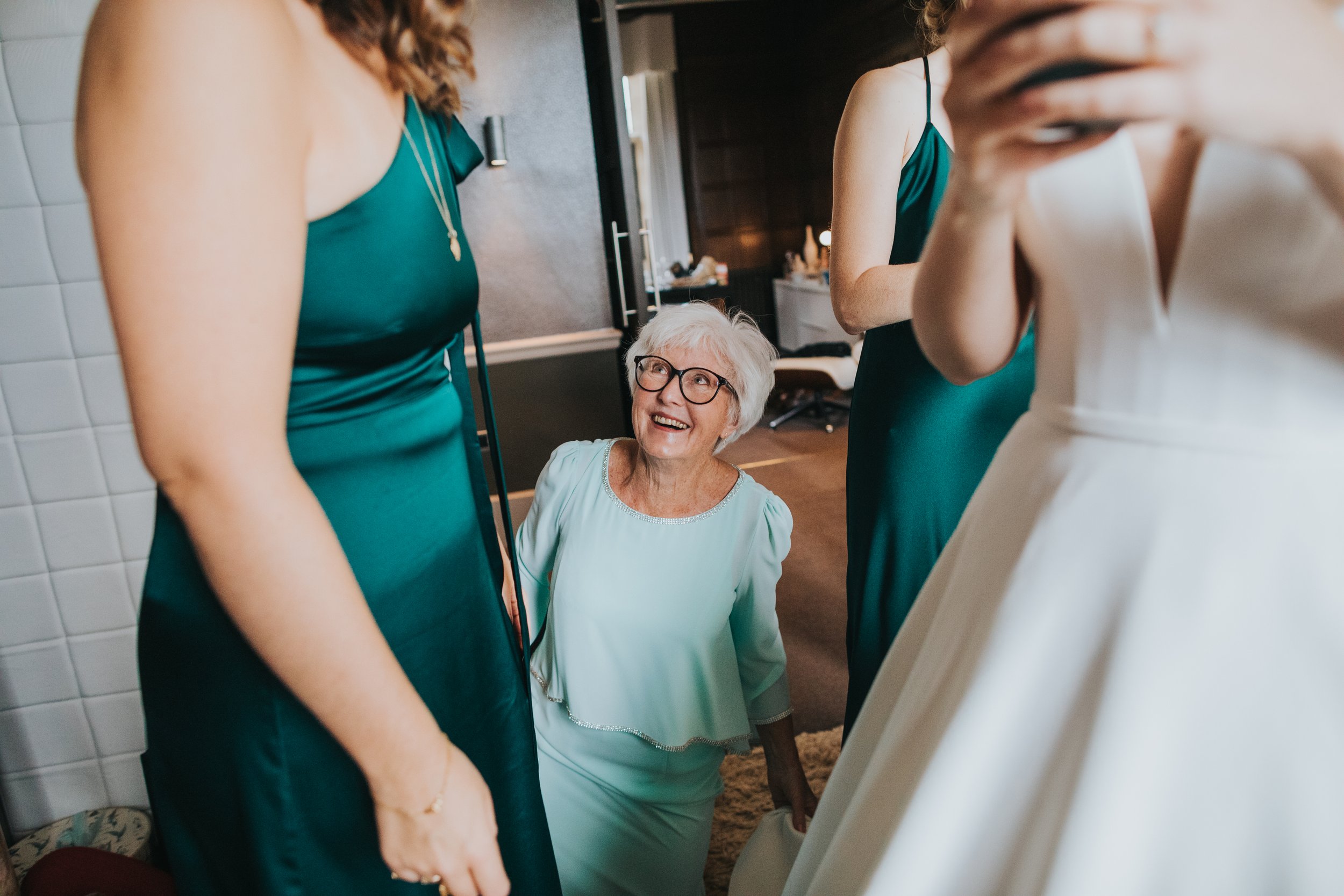 Mother of the Bride kneeling on the floor smiling as she helps her daughter into her dress. 